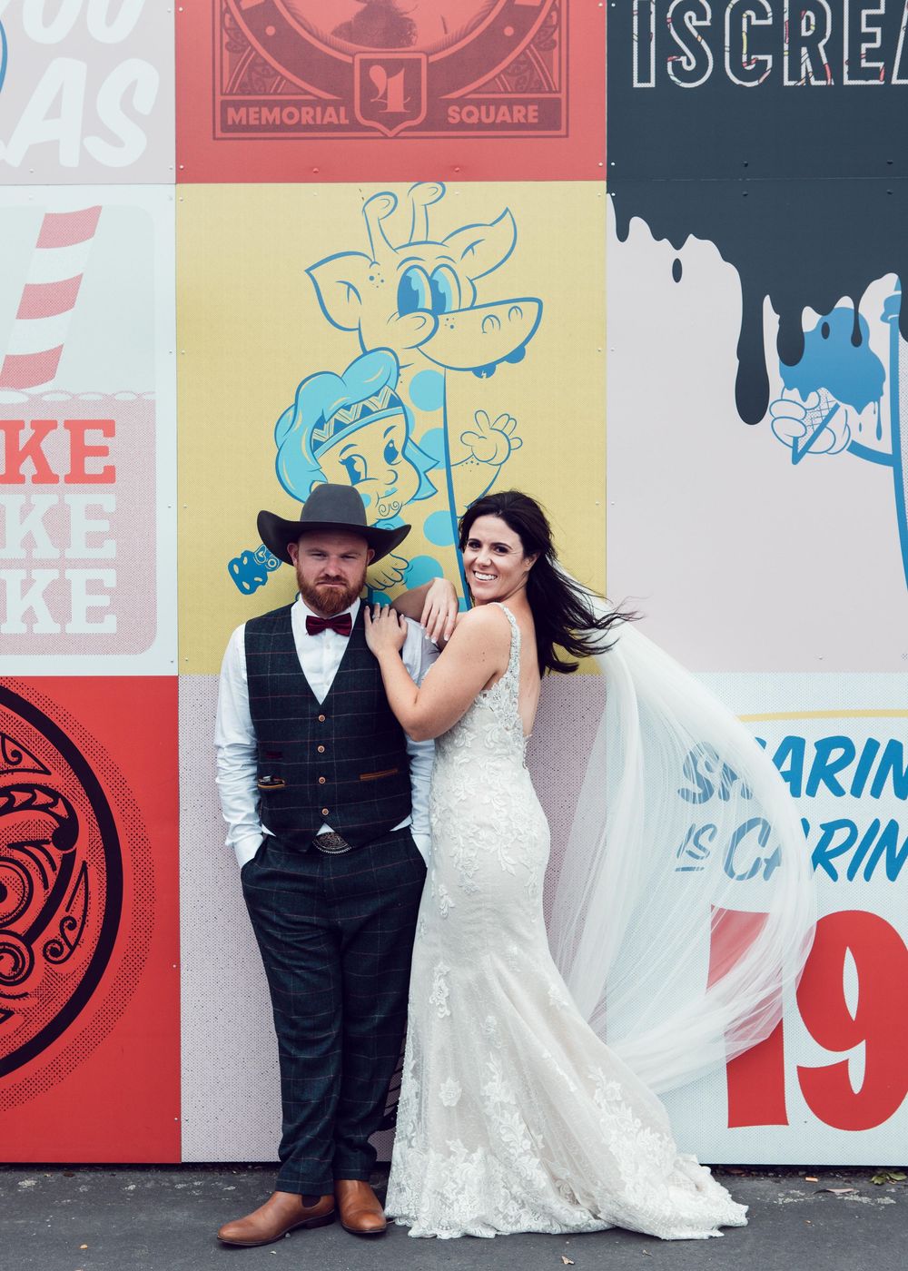 A couple poses in front of a colorful mural wall decorated with cartoon characters and graphics at an outdoor location.