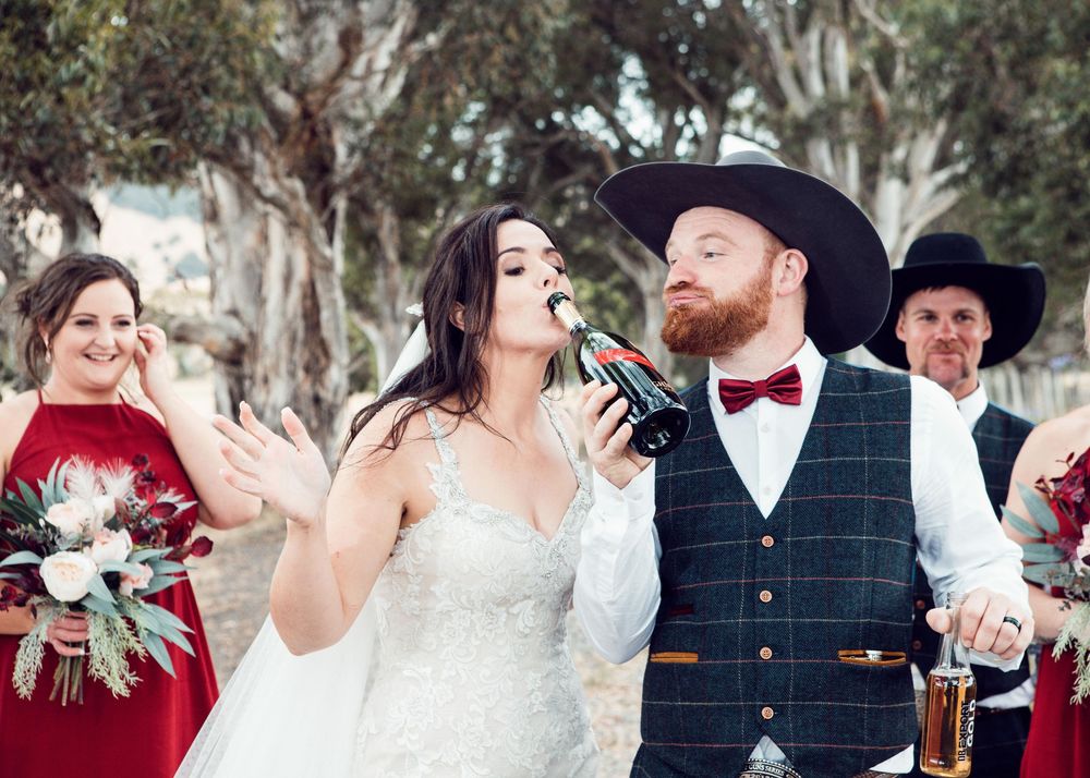 A newlywed couple shares a drink at their rustic outdoor wedding celebration while attendants in red look on.