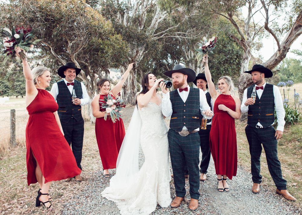 Wedding party in burgundy dresses and black suits celebrating joyfully on a gravel path under trees.