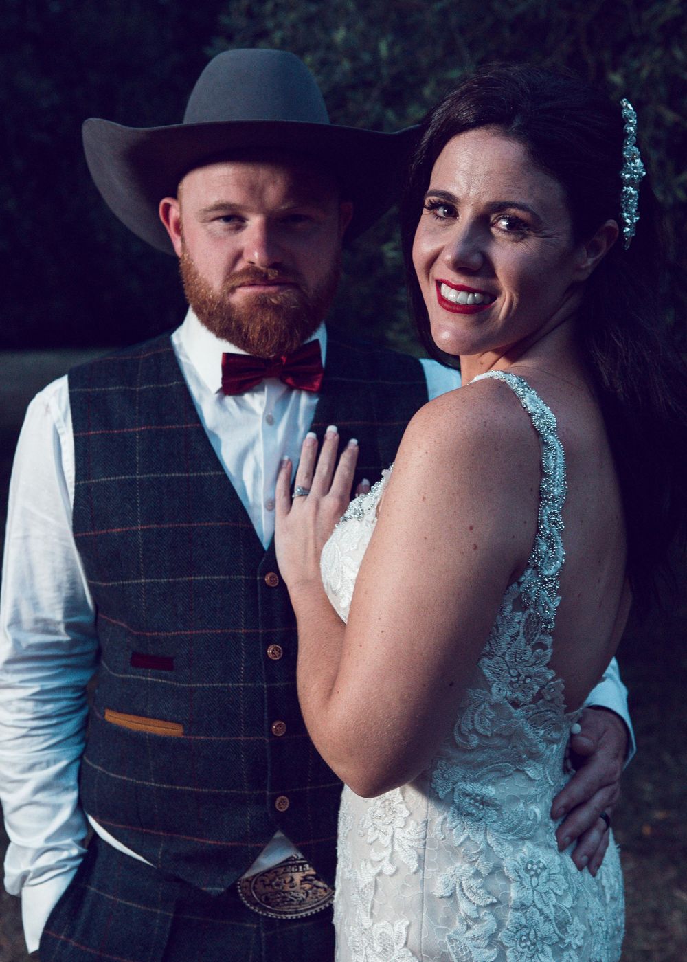 A couple poses together for a romantic wedding photo with the bride in a lace dress and groom wearing a vest and cowboy hat.