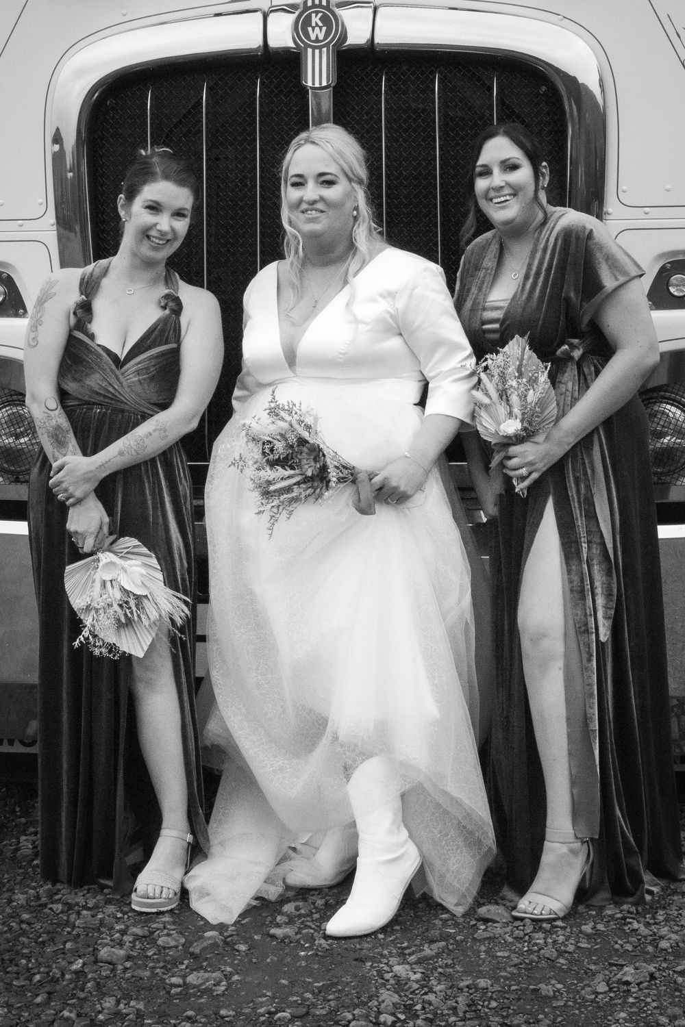 Black and white wedding photo showing a bride in white dress and boots with two bridesmaids in dark dresses outside a vintage car.