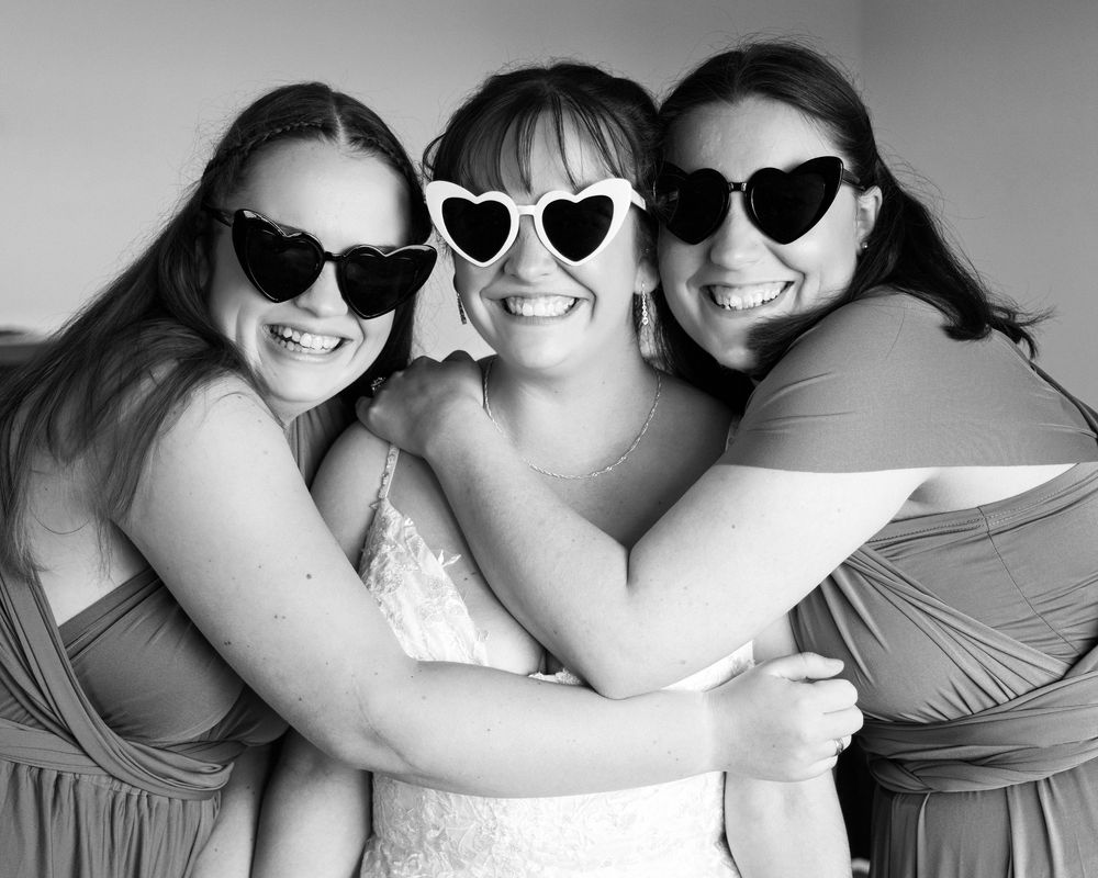 Three friends wearing sunglasses pose together in a black and white photo, with heart-shaped frames and joyful smiles.