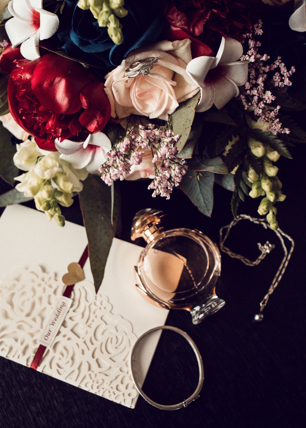 An elegant arrangement of red and pink roses with embossed wedding stationery and delicate jewelry on a dark surface.