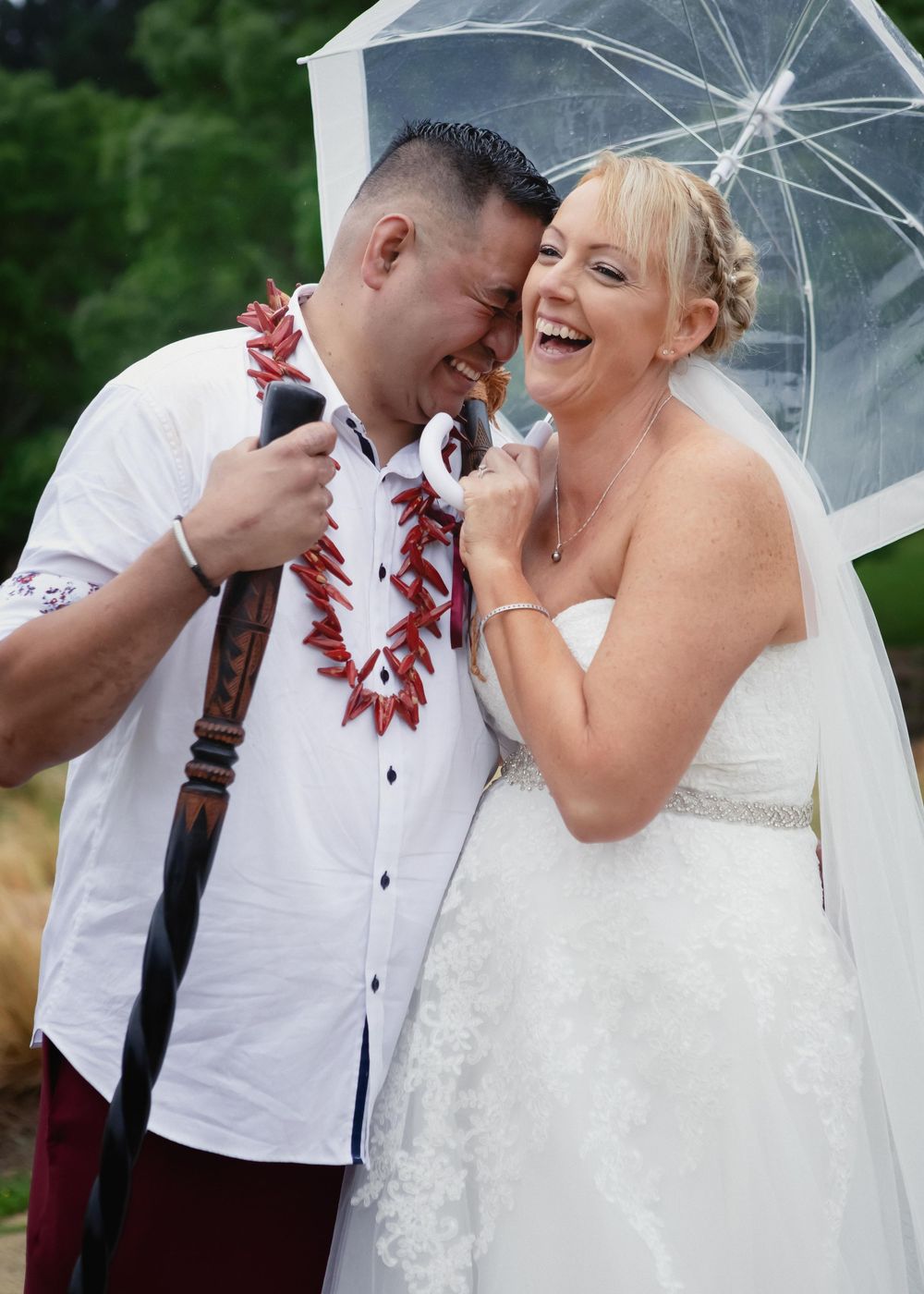 A joyful couple shares a lighthearted moment under a clear umbrella during their wedding photoshoot.