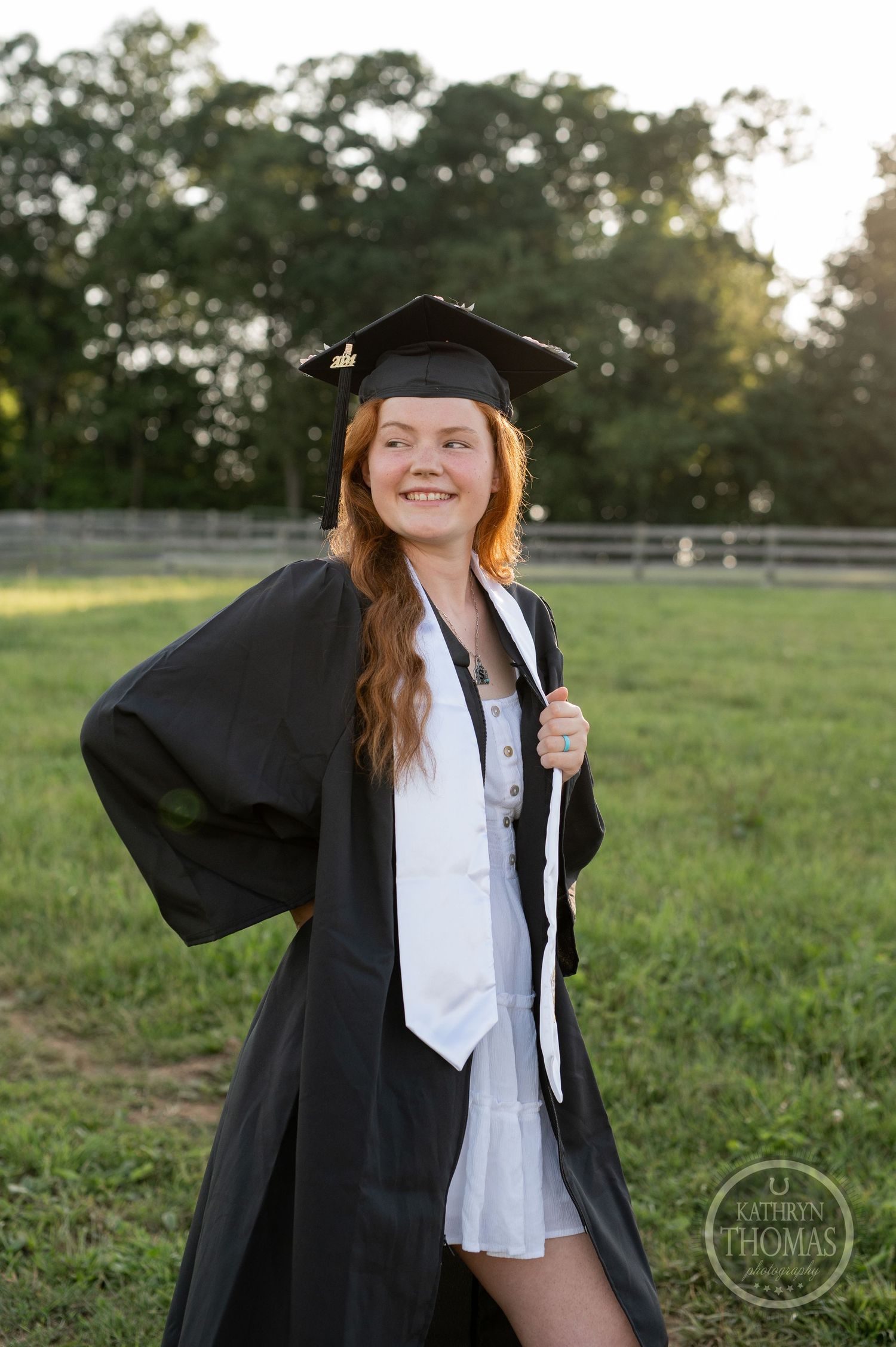 Graduate in black gown and white tie poses in grassy field at sunset.