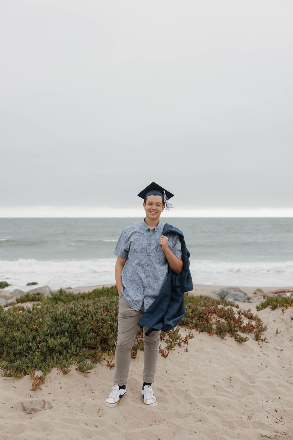 Graduate wearing mortarboard stands on sandy beach with ocean and cloudy sky in background.