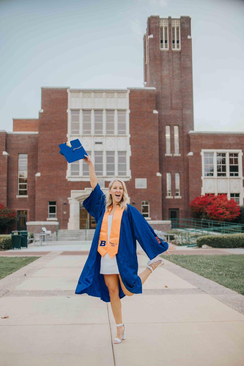 Person in blue graduation gown celebrates on campus path in front of traditional brick university building with tower.