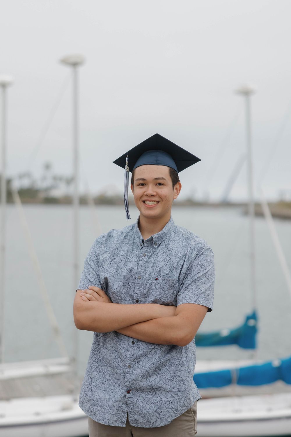 Graduate in blue patterned shirt and graduation cap poses with crossed arms at a marina.