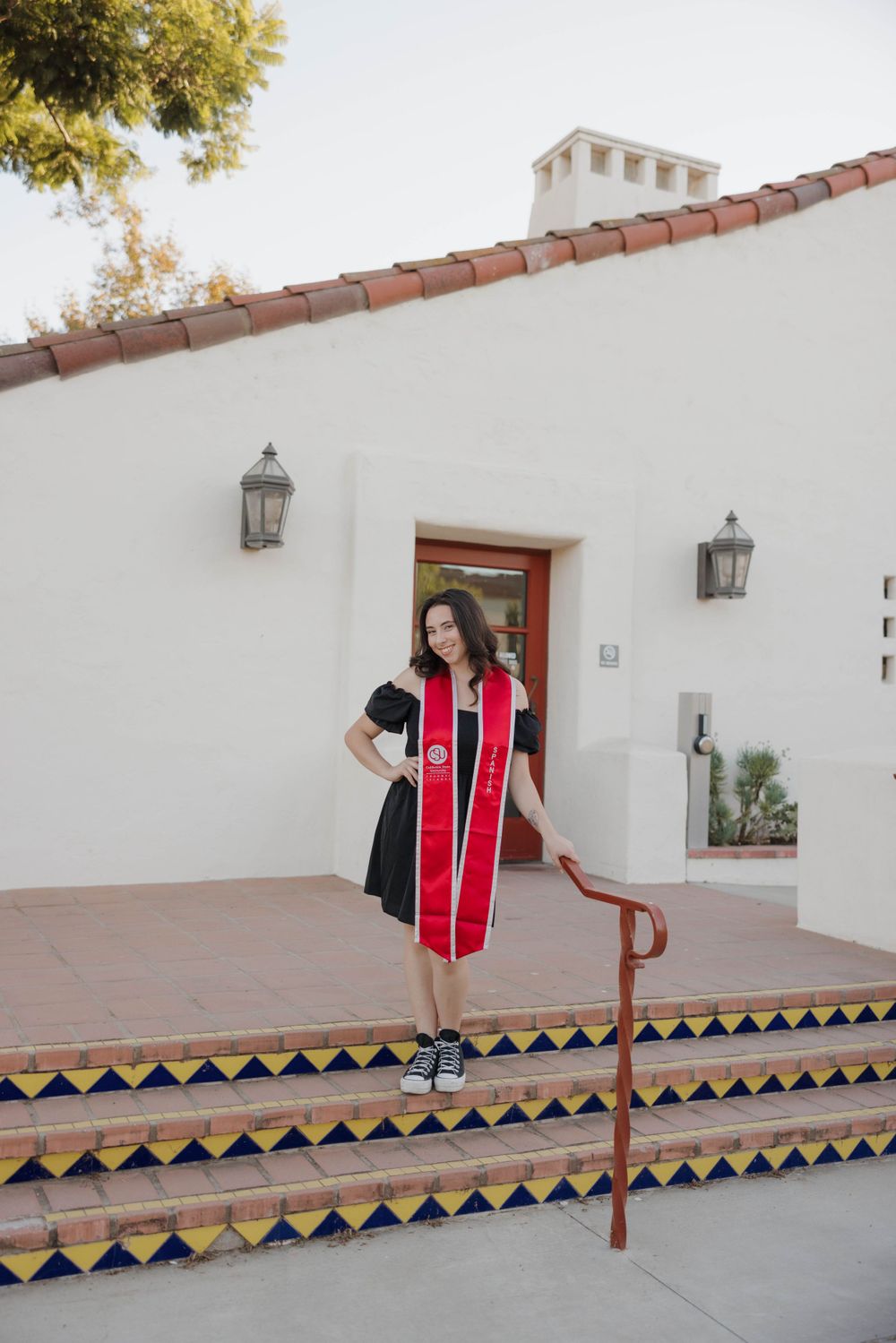 Graduate wearing a black and red graduation gown poses on decorative tiled steps outside a white Spanish-style building.
