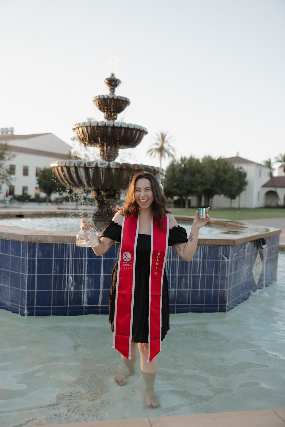 Graduate in red and black graduation stole celebrates in front of fountain at university campus.