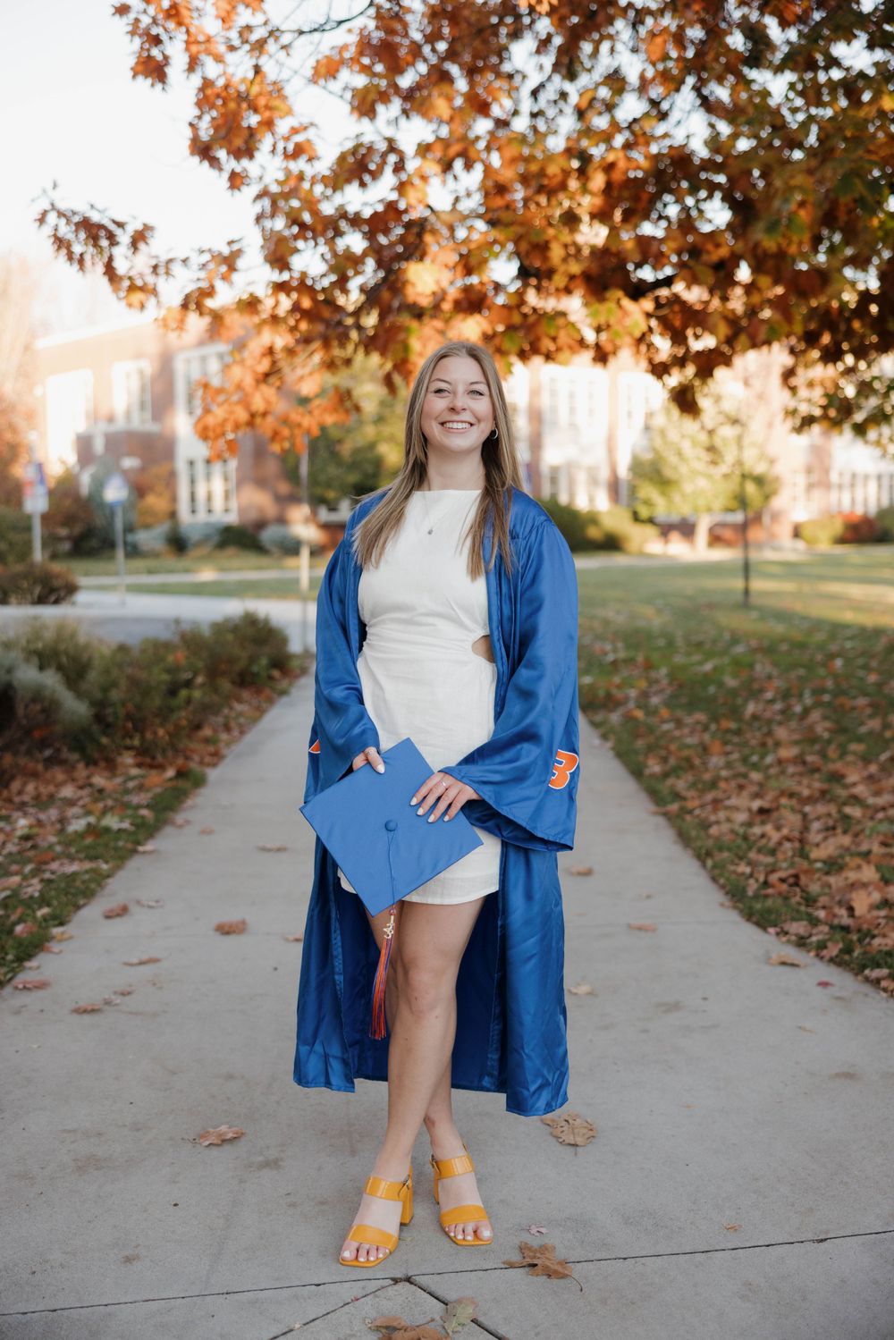 A graduate in a blue cap and gown stands on a sidewalk surrounded by autumn leaves and yellow sandals on a sunny day.