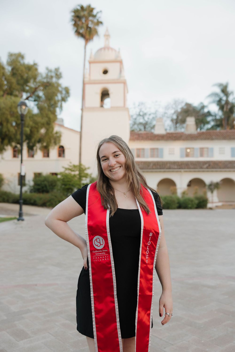 Graduate wearing red graduation stole smiles in front of Spanish colonial style church with bell tower and palm trees.