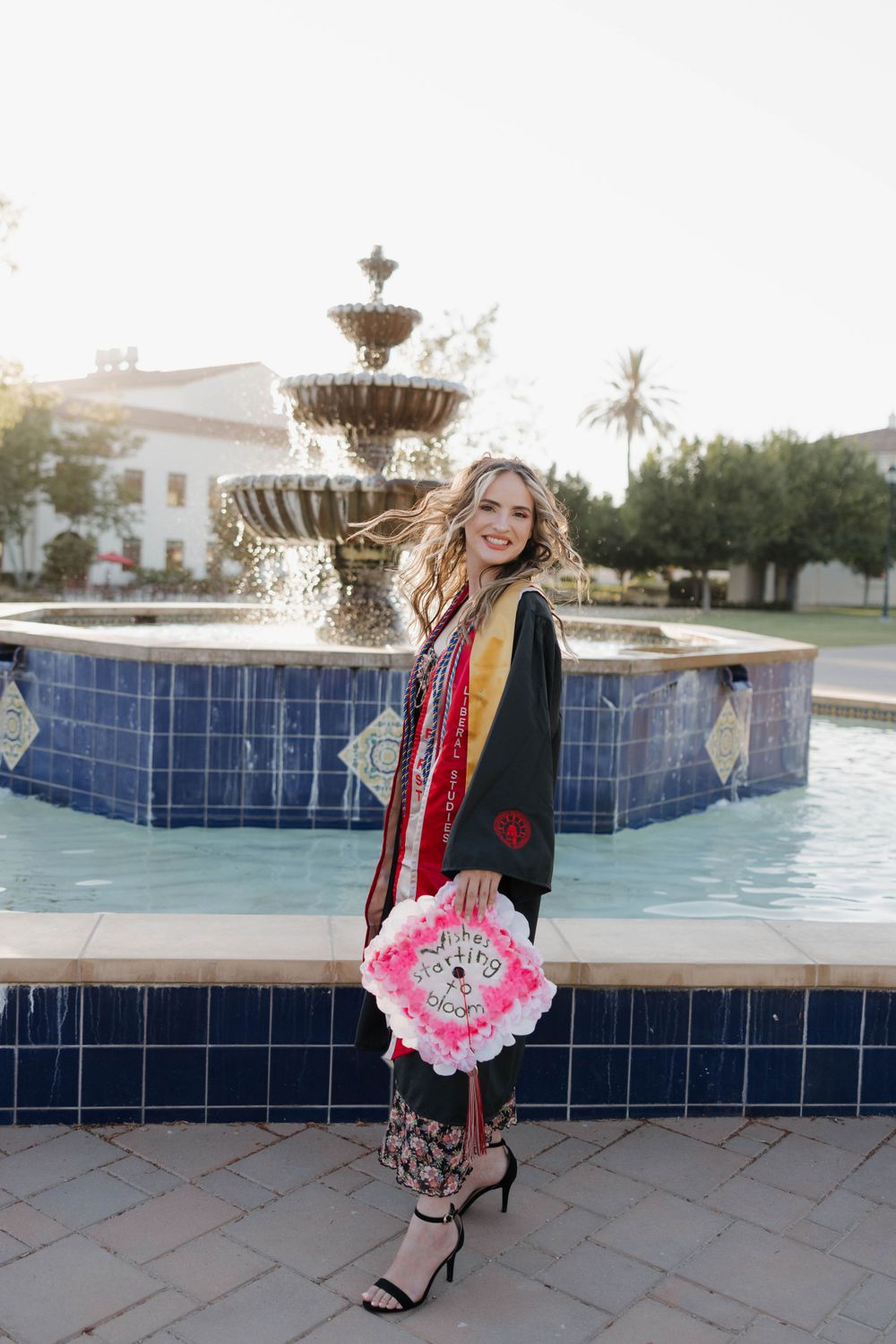 Graduate in cap and gown poses with decorated graduation cap in front of ornate tiered fountain at university campus.