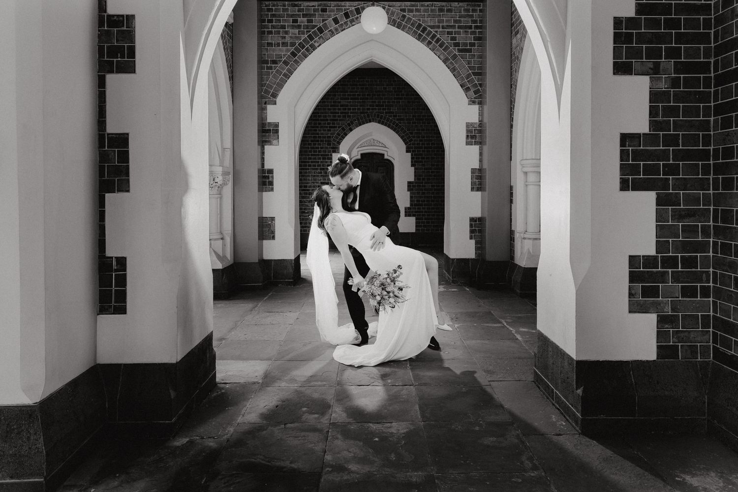 A black and white photo of a couple dancing in an arched church hallway, captured by Apertured Media Auckland Wedding Photographer.