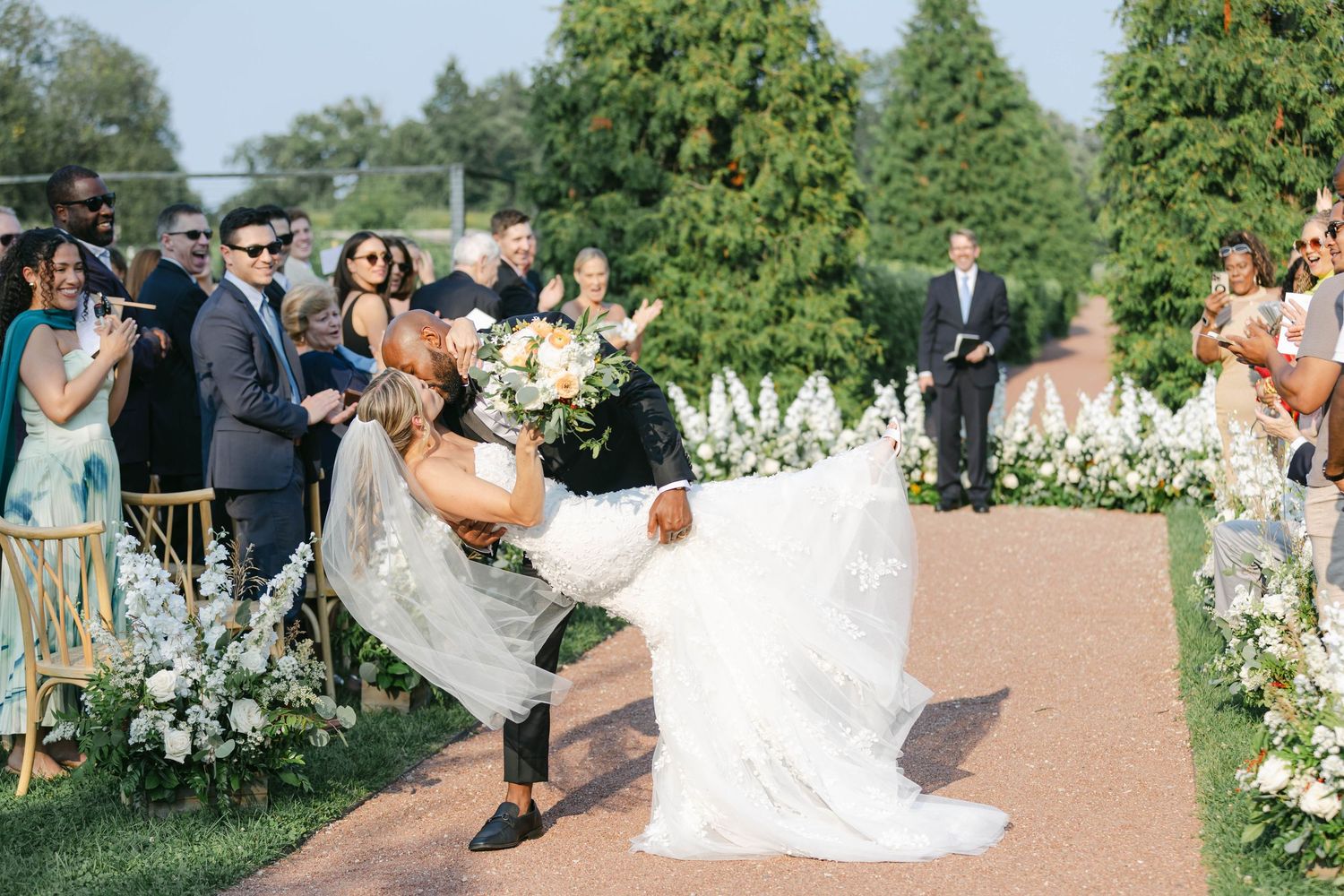 A wedding couple dances with guests at an outdoor garden reception surrounded by lush greenery and white chairs. - Elawa Farm