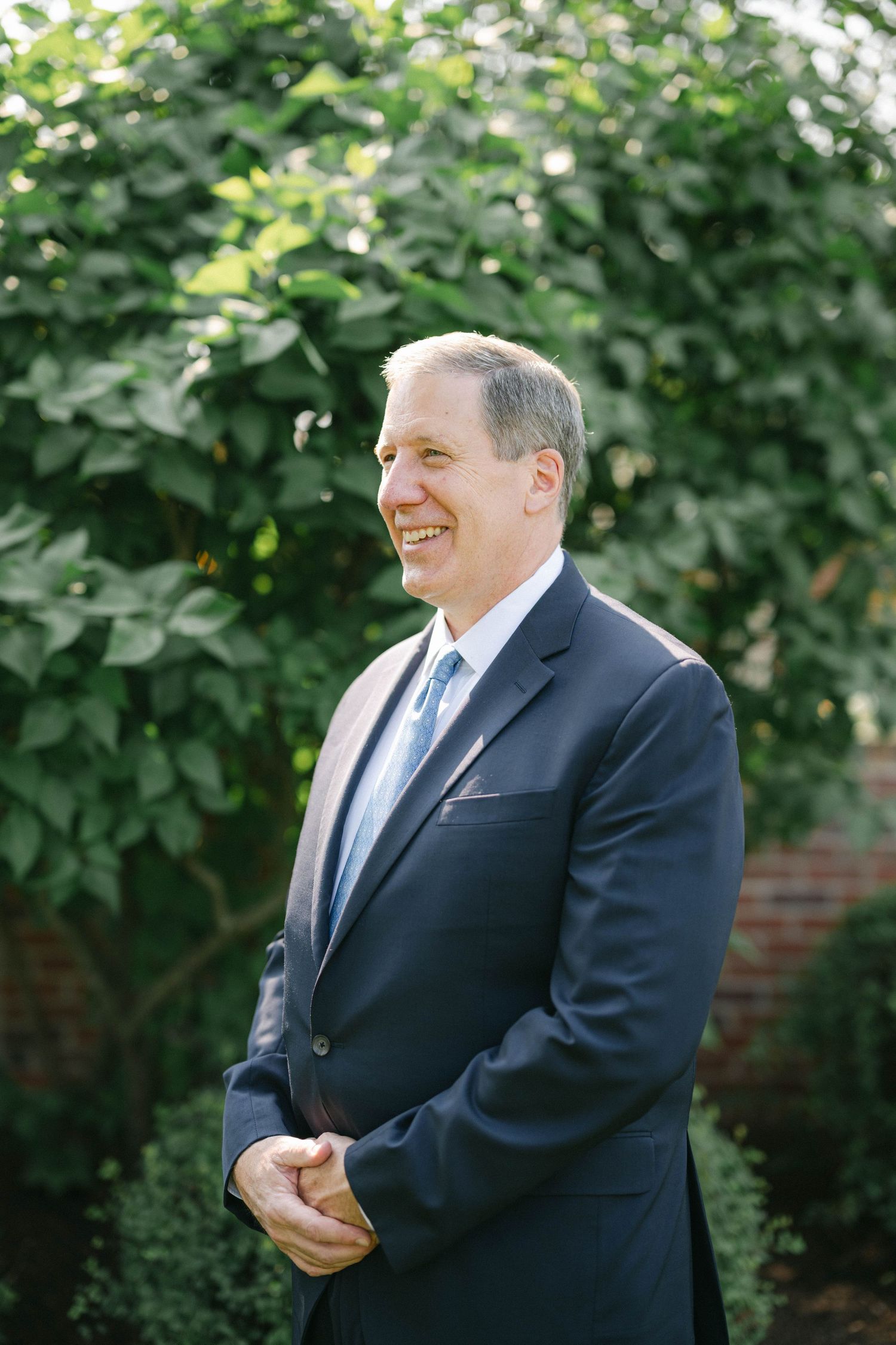 A person in a navy suit and blue tie smiles while standing outdoors near green foliage. - Elawa Farm