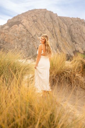 Woman in flowing white dress stands among tall beach grass with mountain backdrop.