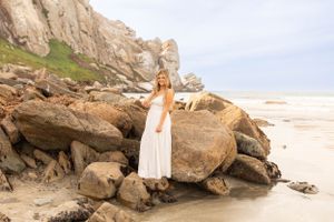 Woman in white dress stands on coastal rocks with dramatic cliffside backdrop.