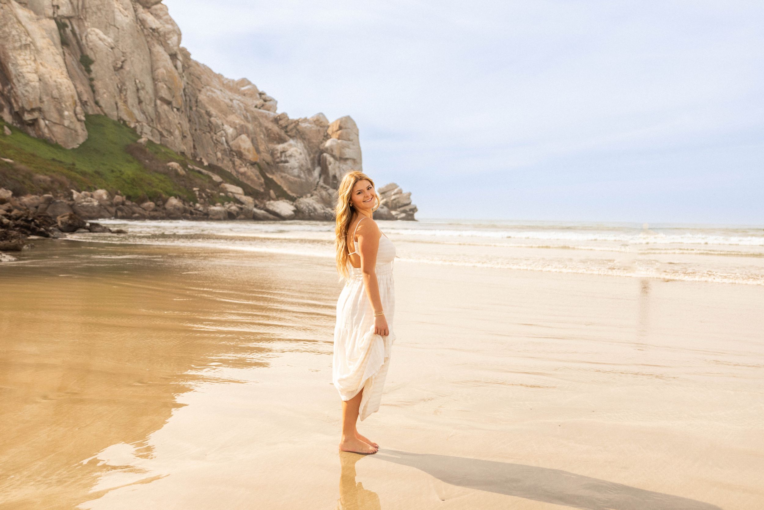 Woman in white dress walks along shoreline with dramatic coastal cliffs in background.