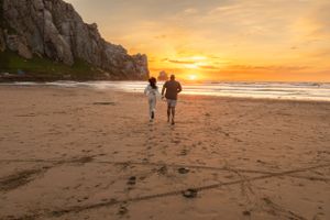 Couple walking along empty beach at sunset with cliffs and footprints in sand.