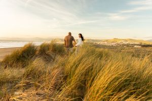 Couple walking through tall beach grass with ocean and sunset in background.