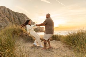 Couple dancing playfully on sandy beach path with dramatic cliff and sunset view.