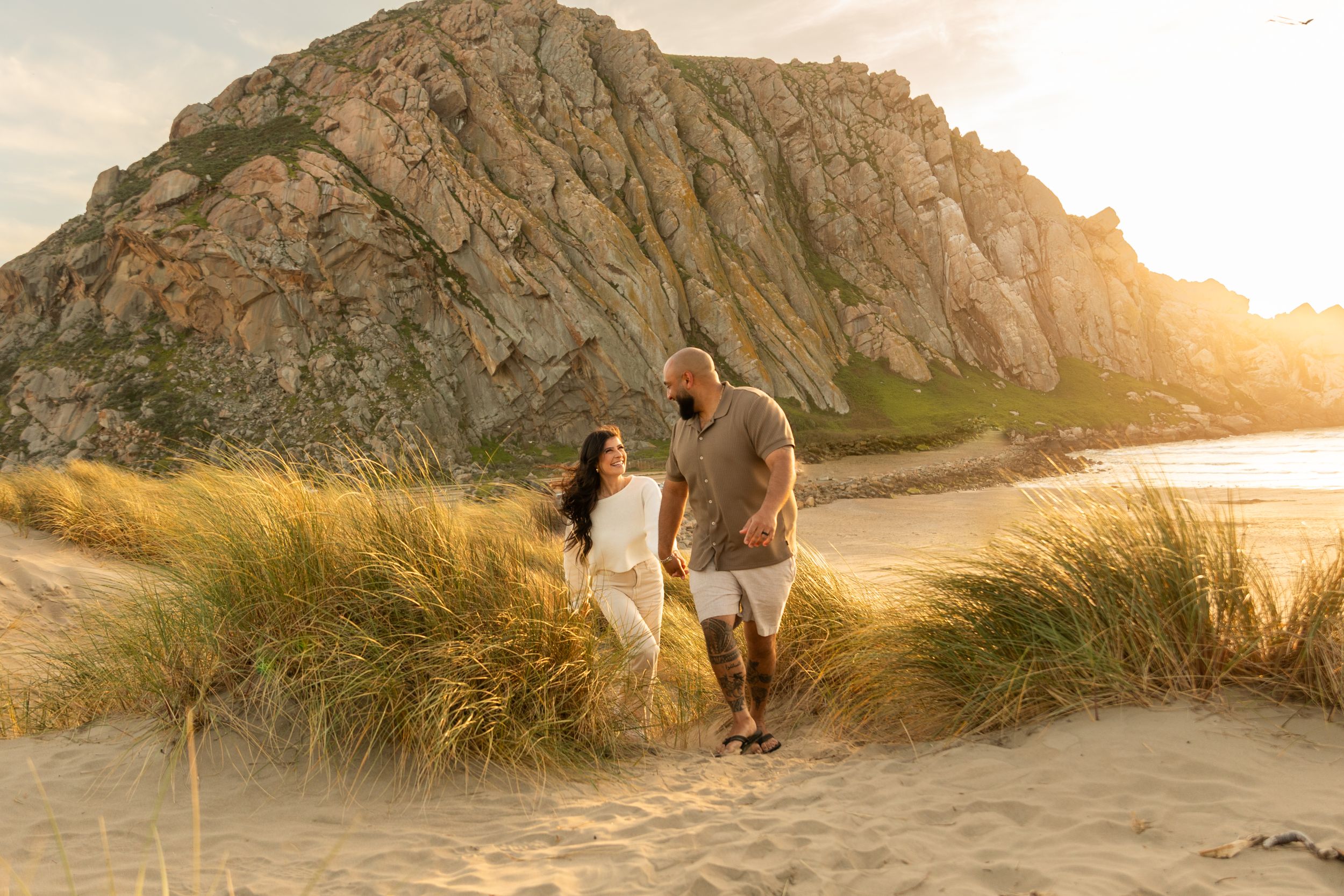 Couple walking together on beach path with dramatic rocky cliff and sunset backdrop.