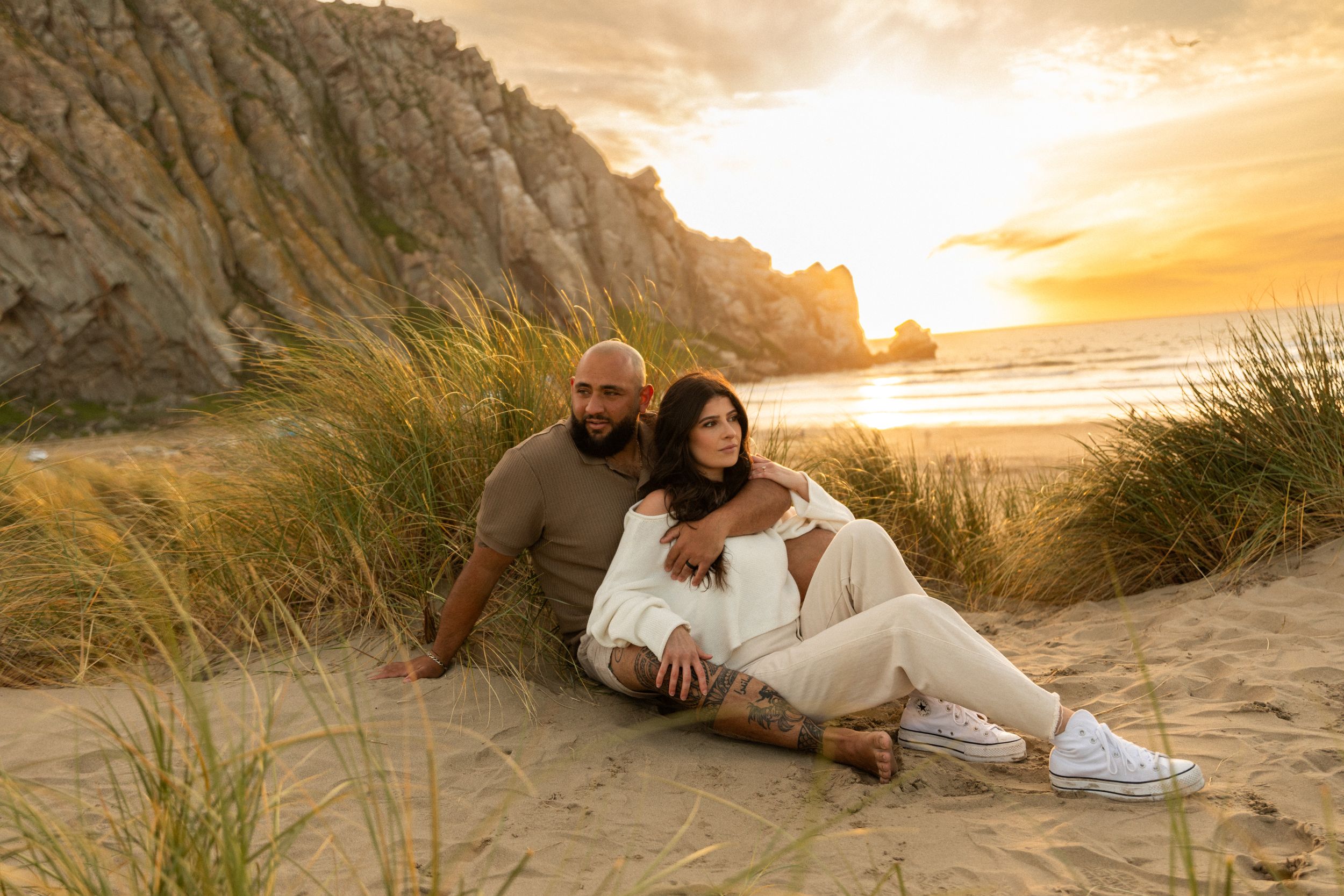 Couple relaxes on sandy beach against rocky coastline during golden sunset.