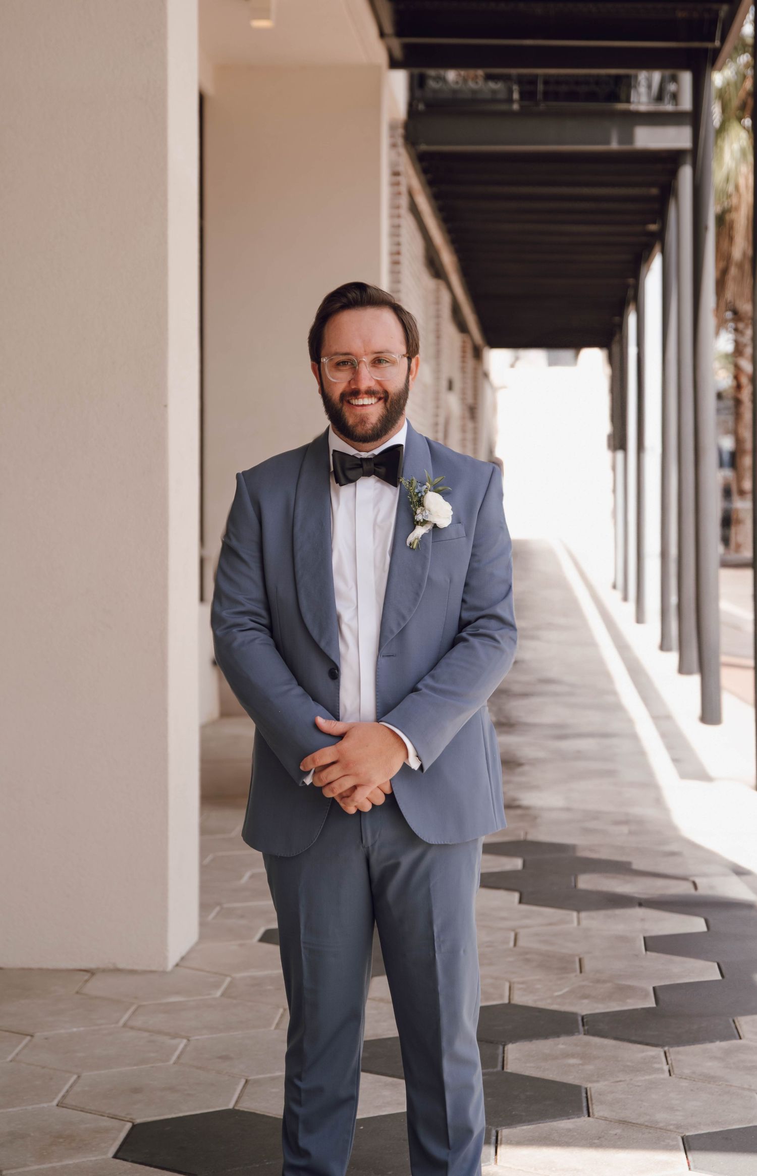 A person in a blue-gray suit and white boutonniere poses in an elegant covered walkway with columns.