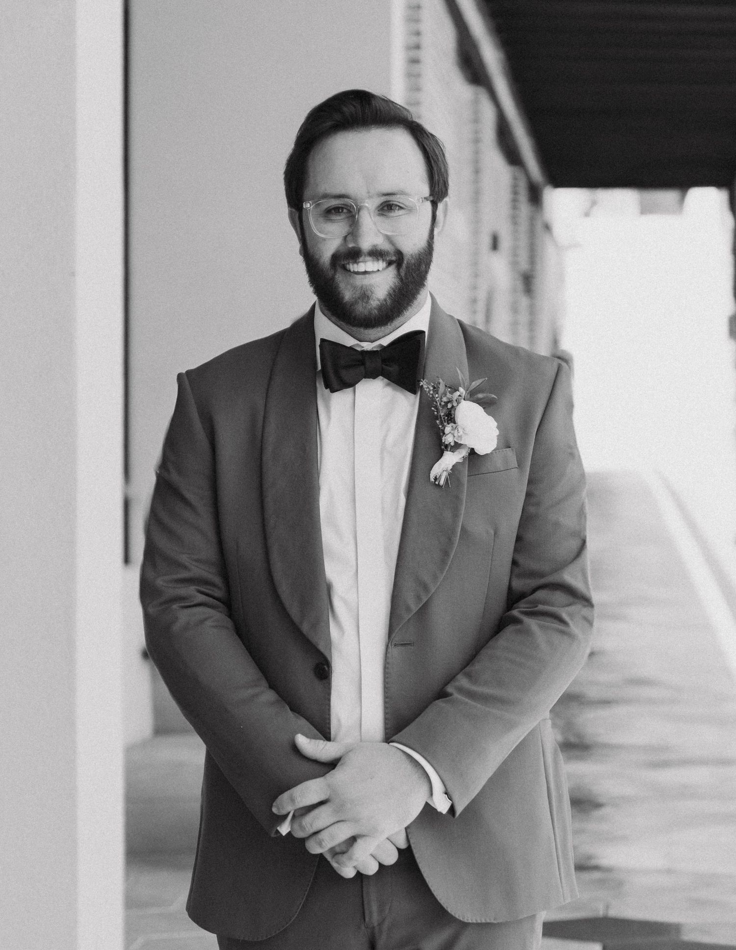 A groom in a dark suit and bowtie poses for formal wedding portraits in a bright hallway.