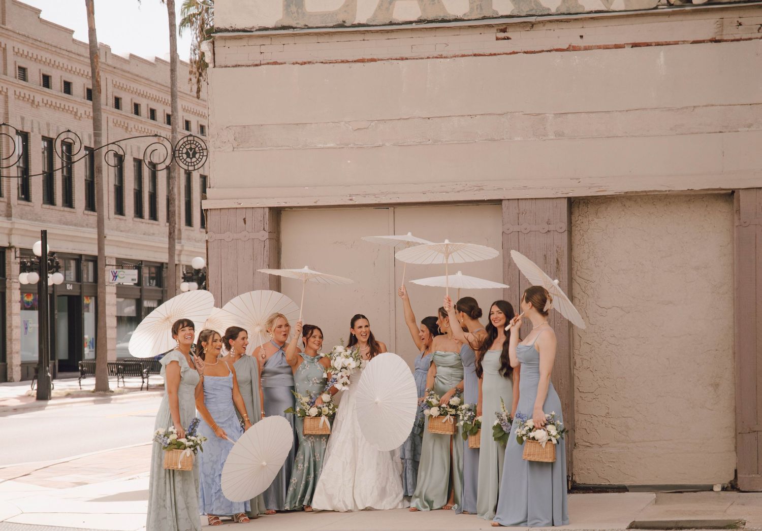 A wedding party in light blue bridesmaid dresses holding white umbrellas poses together on a sunny street.