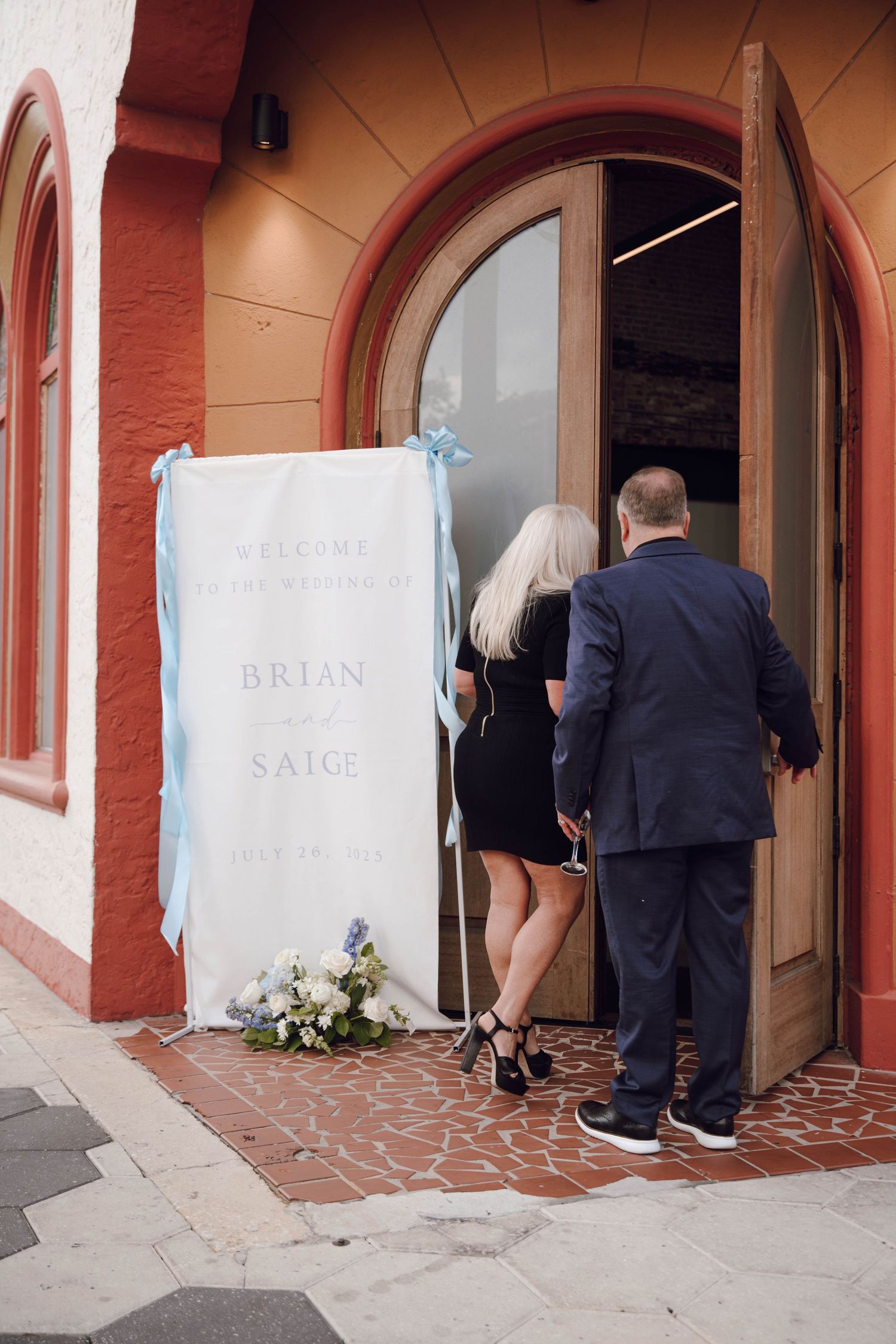Two people entering a doorway with an arched entrance and a white banner with text displayed outside.