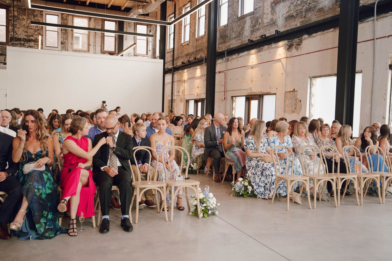 Wedding guests seated in rows of wooden chairs in an industrial-style venue with exposed brick walls.