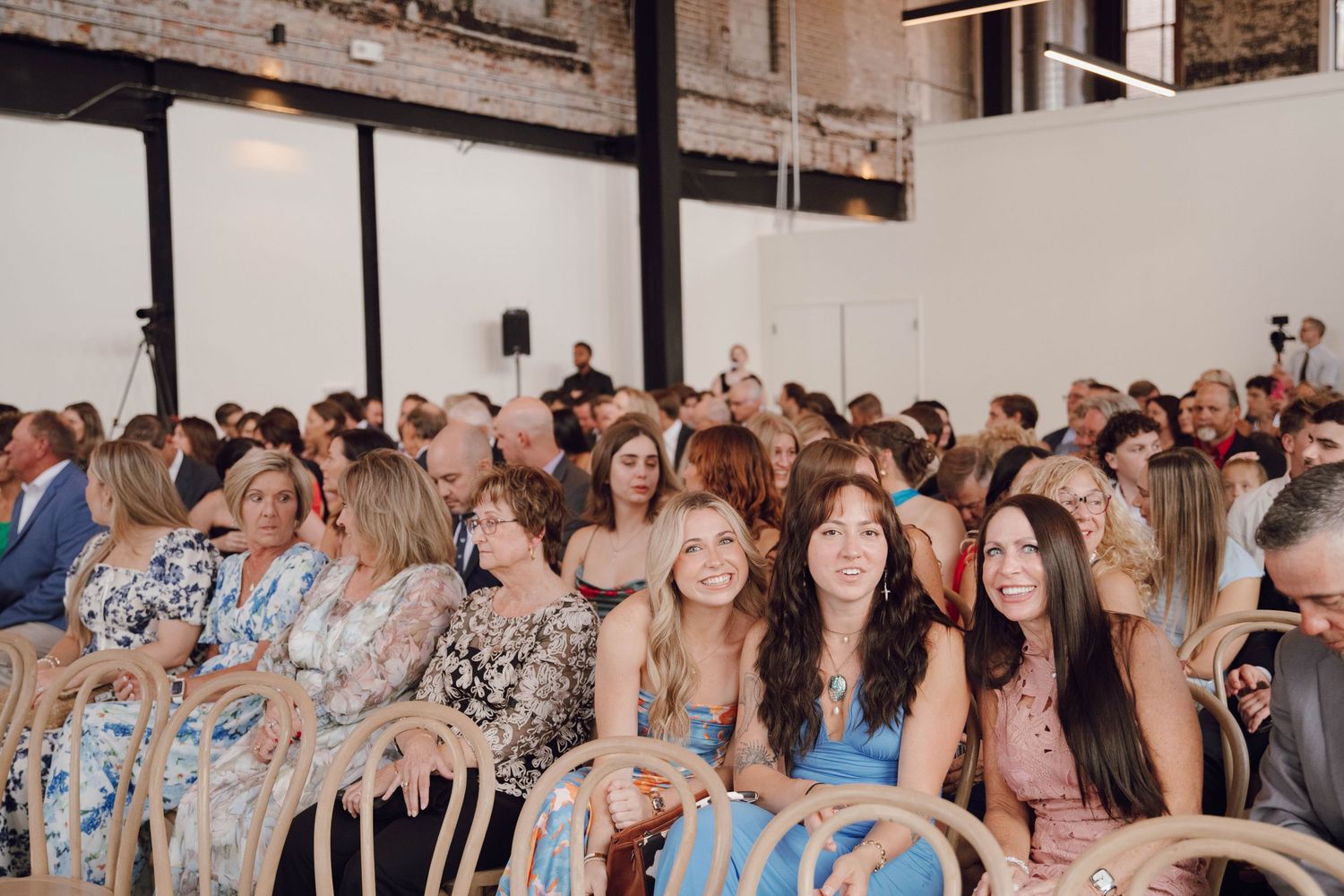Wedding guests seated in rows at an indoor venue with exposed brick walls and natural lighting.