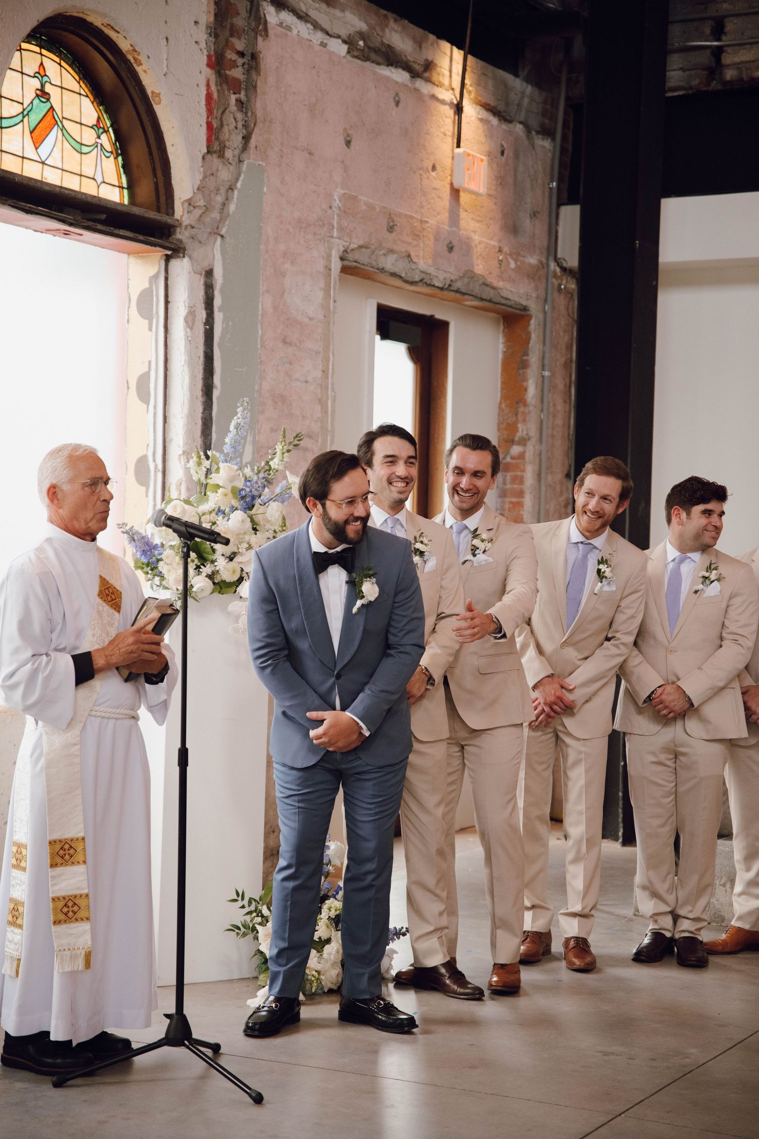 Groomsmen in tan and gray suits lined up against a rustic brick wall during wedding ceremony.
