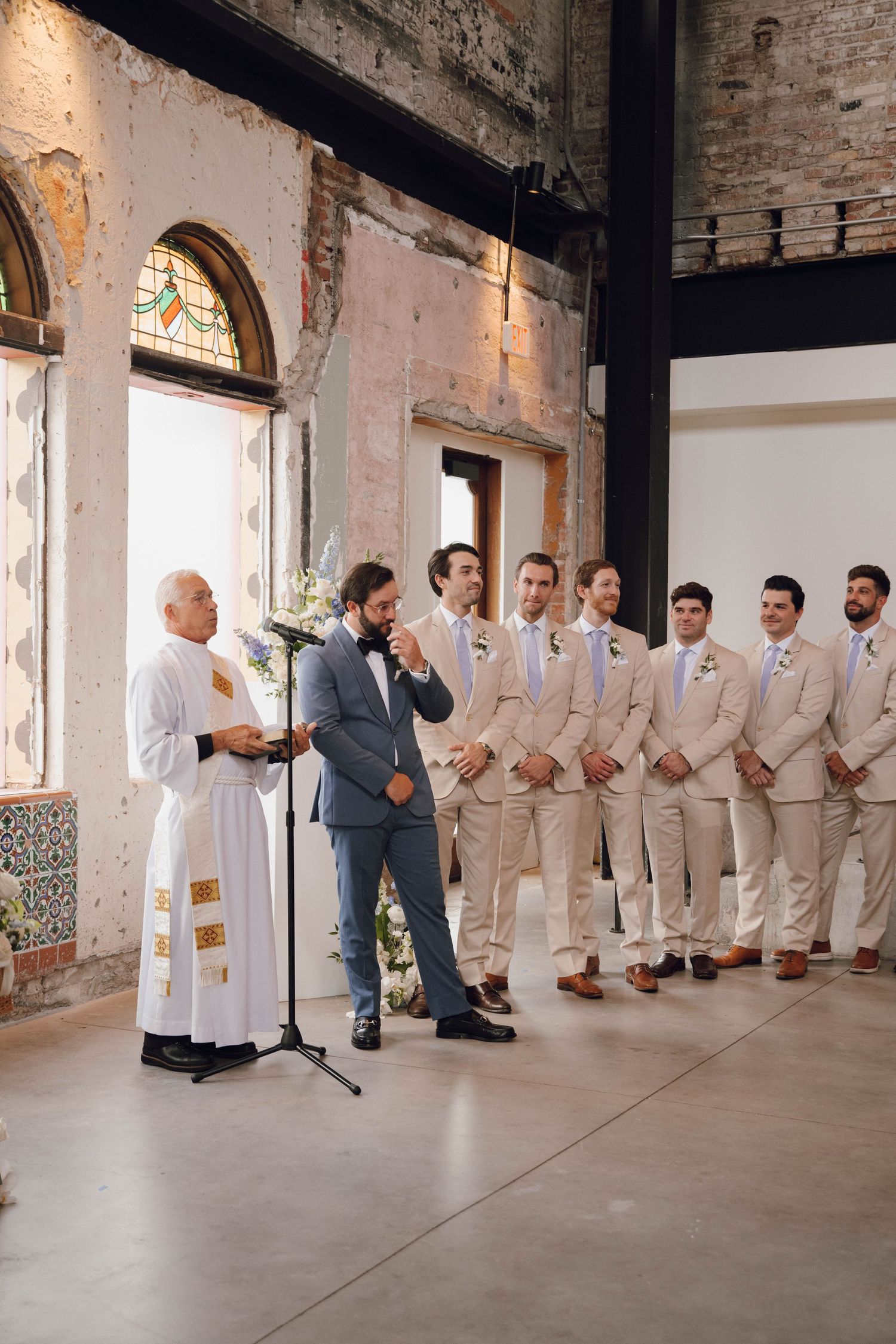 Wedding ceremony in a rustic brick venue with groomsmen in beige suits lined up beside the officiant.