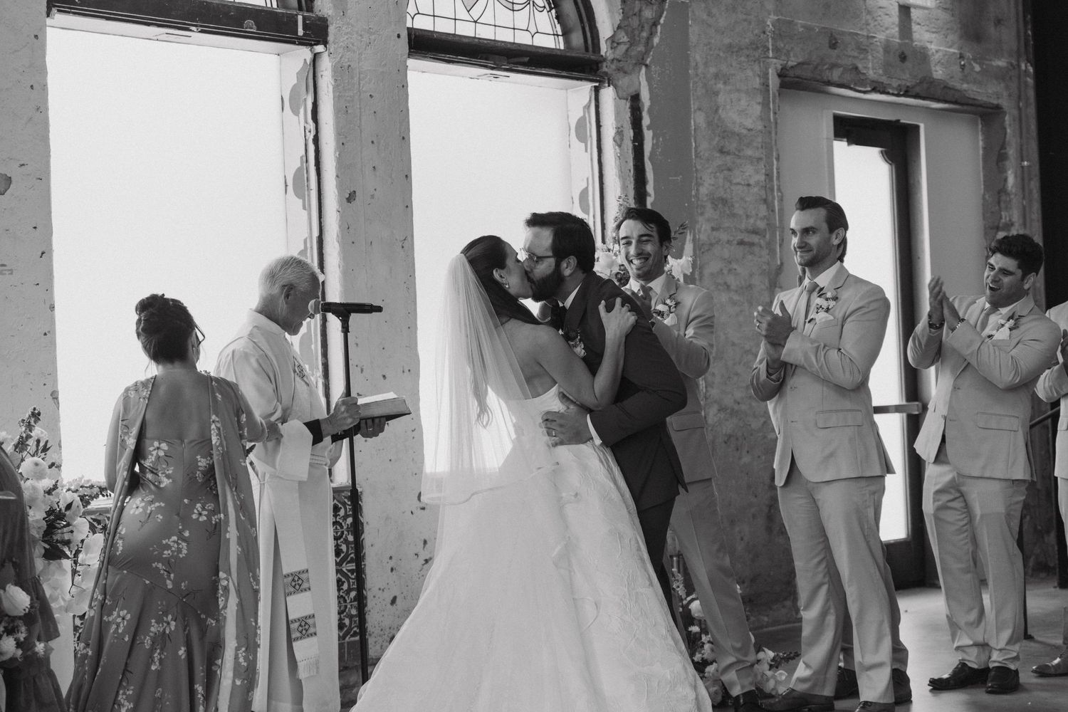 Wedding guests gather in an industrial venue with exposed brick walls and large windows.