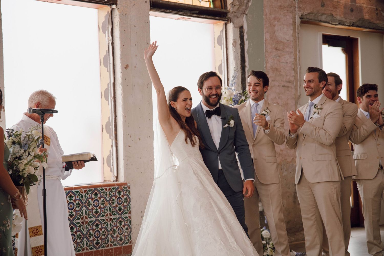 Wedding party in formal attire poses together in a rustic brick venue with arched windows.