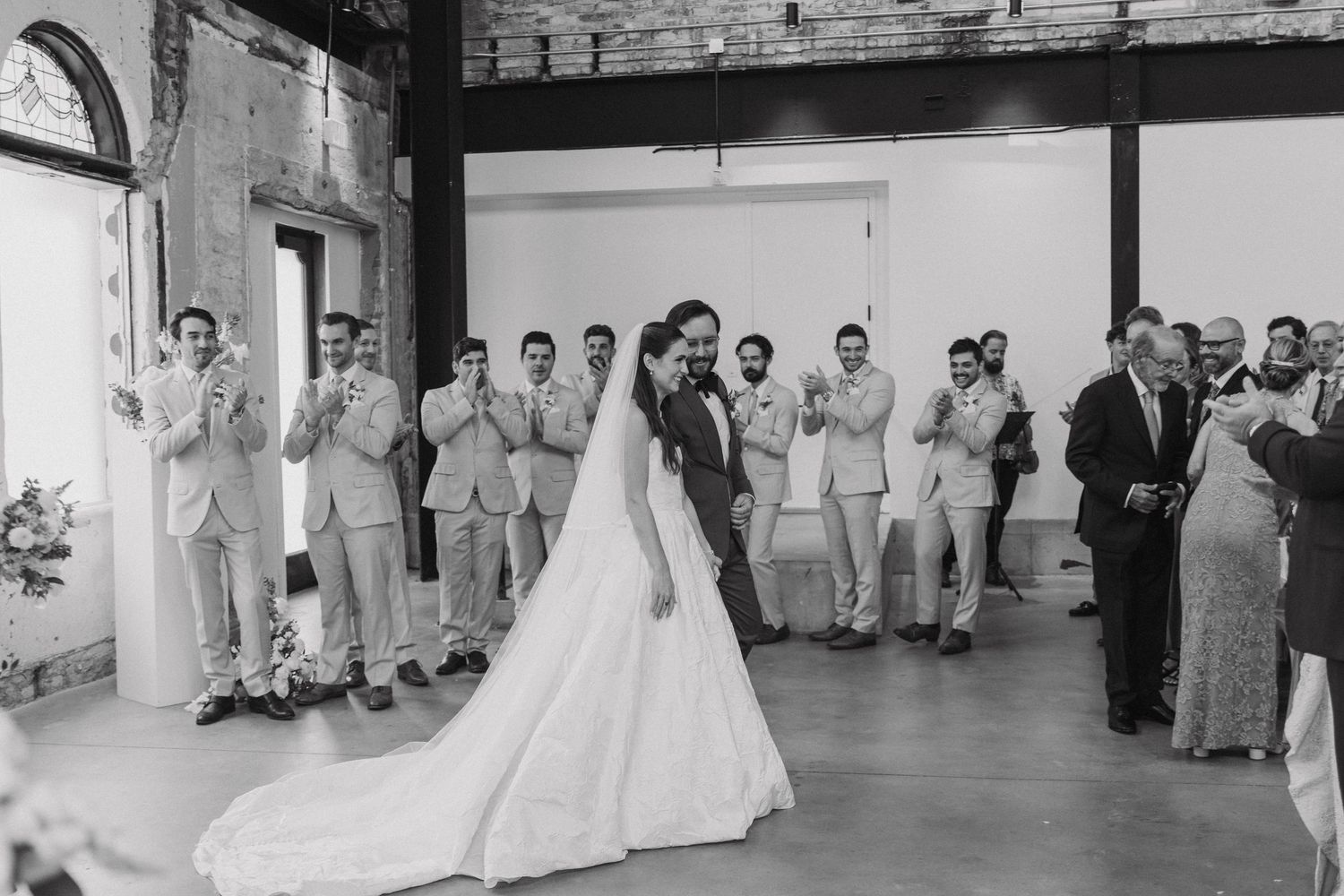 Wedding party lined up in formal attire against an industrial brick wall backdrop.