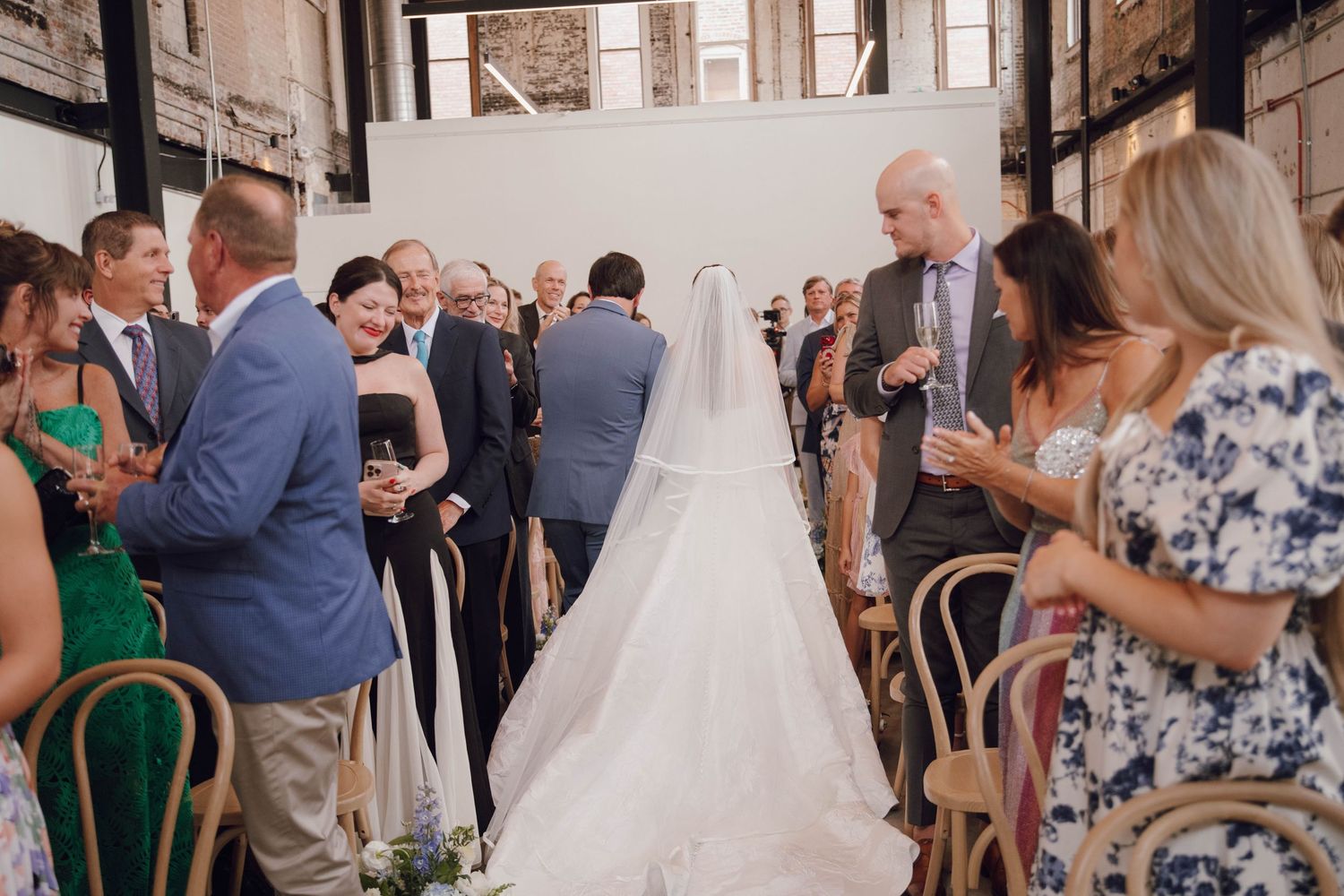 Wedding guests gather in a rustic indoor venue with exposed brick walls and wooden chairs.