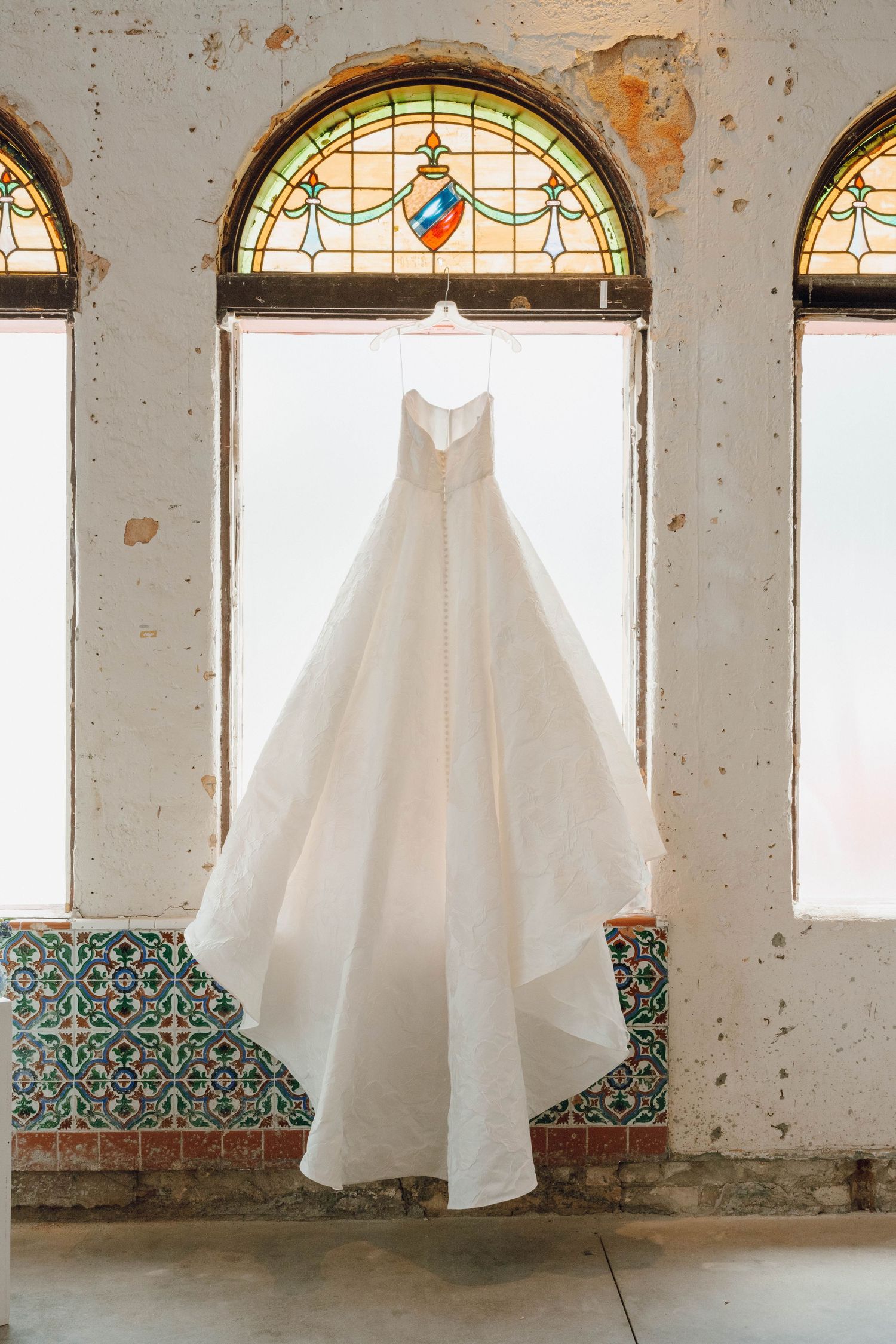 A white wedding dress hangs in an abandoned room with stained glass windows and decorative tiled walls.