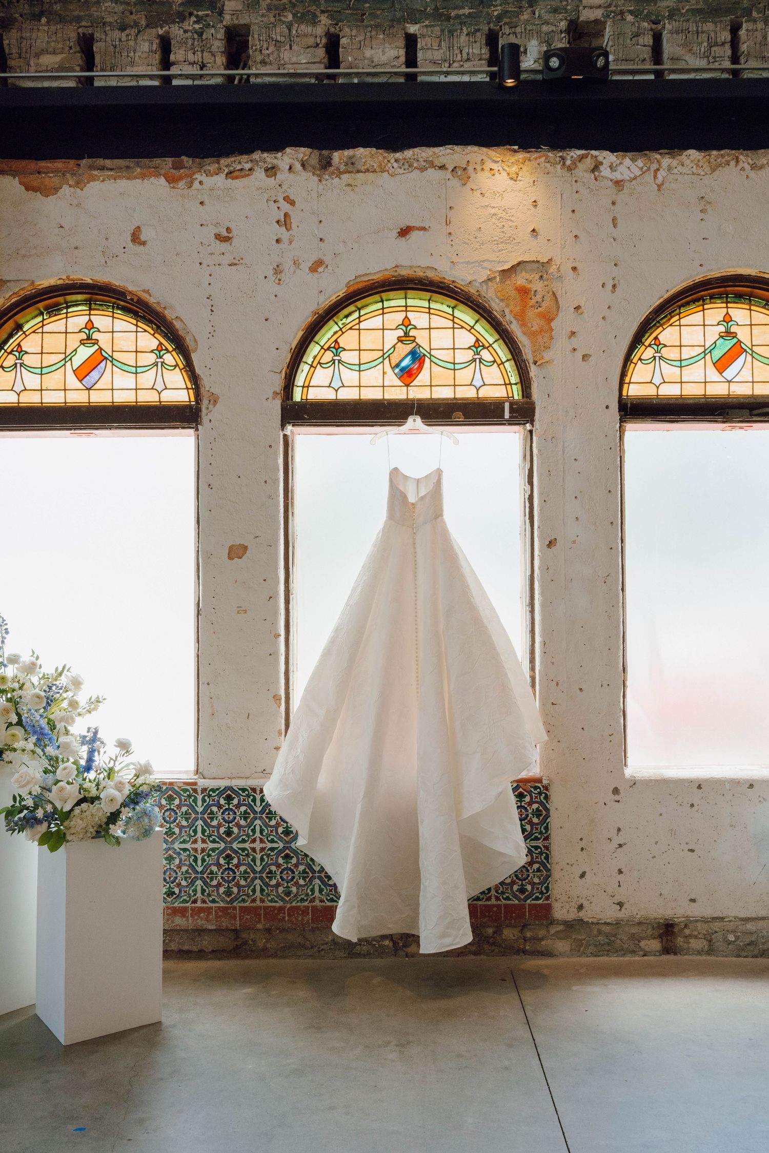An elegant white wedding dress hangs in an archway with stained glass windows and decorative tile details.