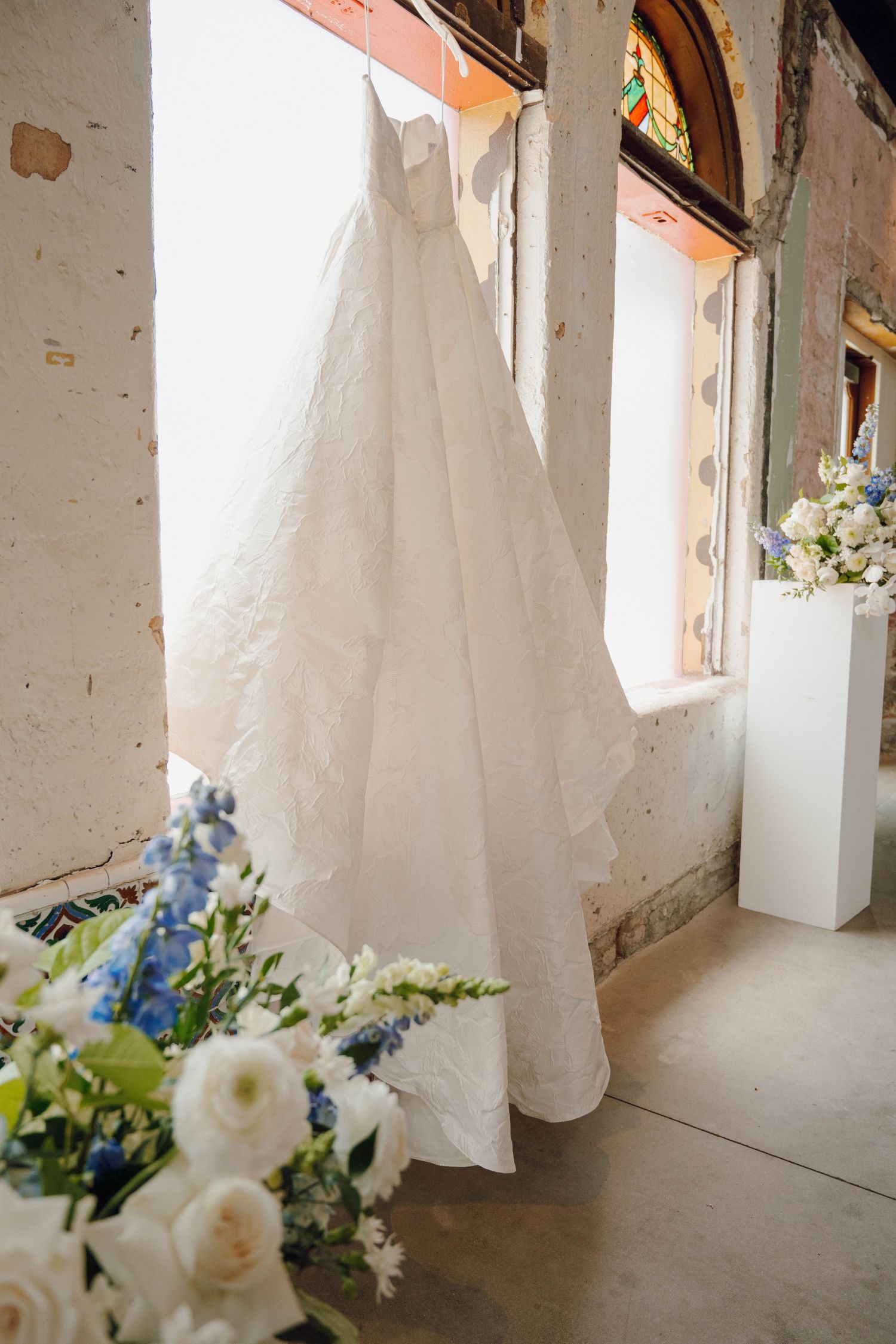 A white bridal gown is displayed against arched windows with floral arrangements of roses and delphinium nearby.