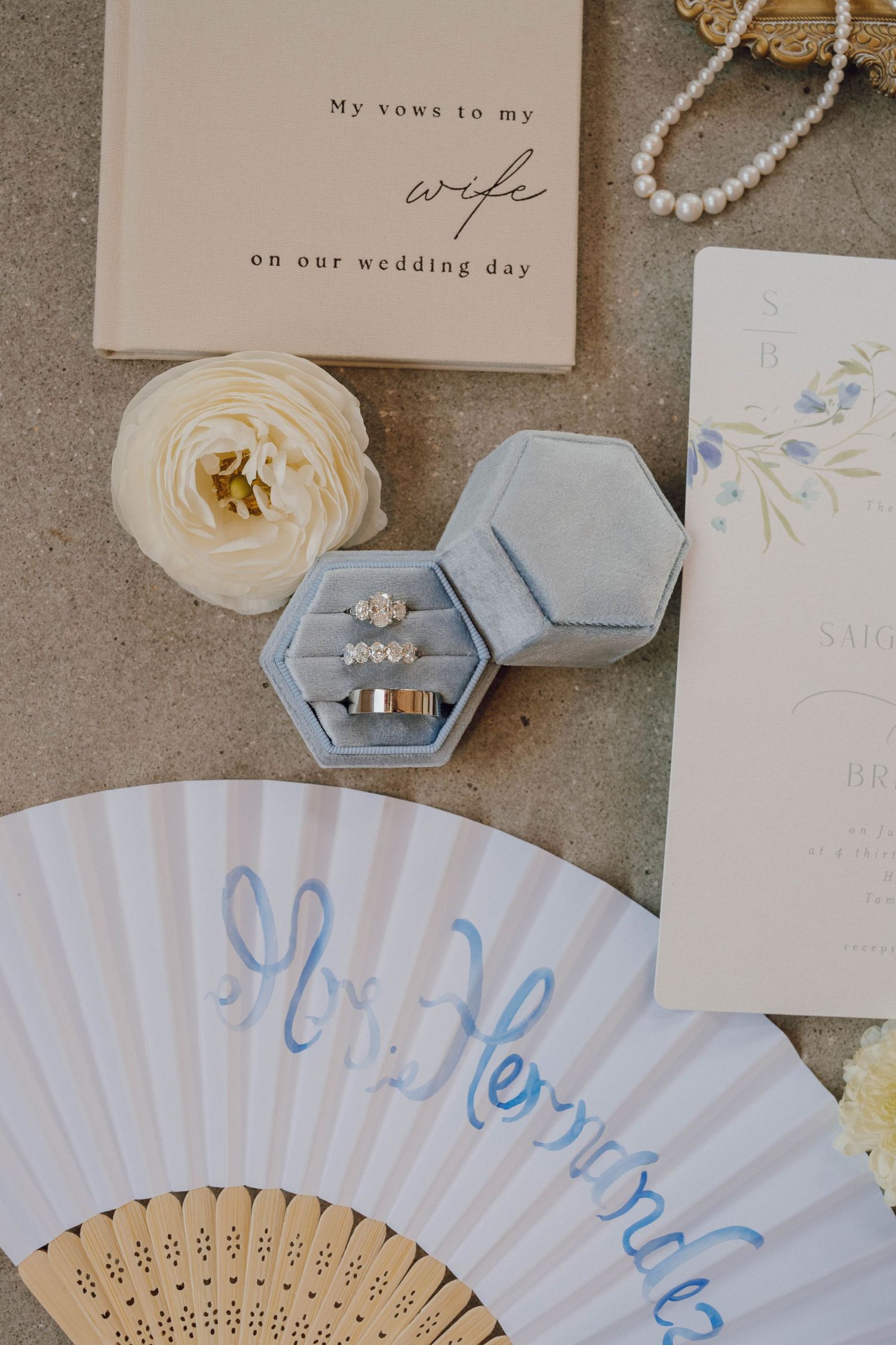 Wedding details flatlay with paper fans, rings, vow books, and delicate floral stationery in soft blue and white tones.