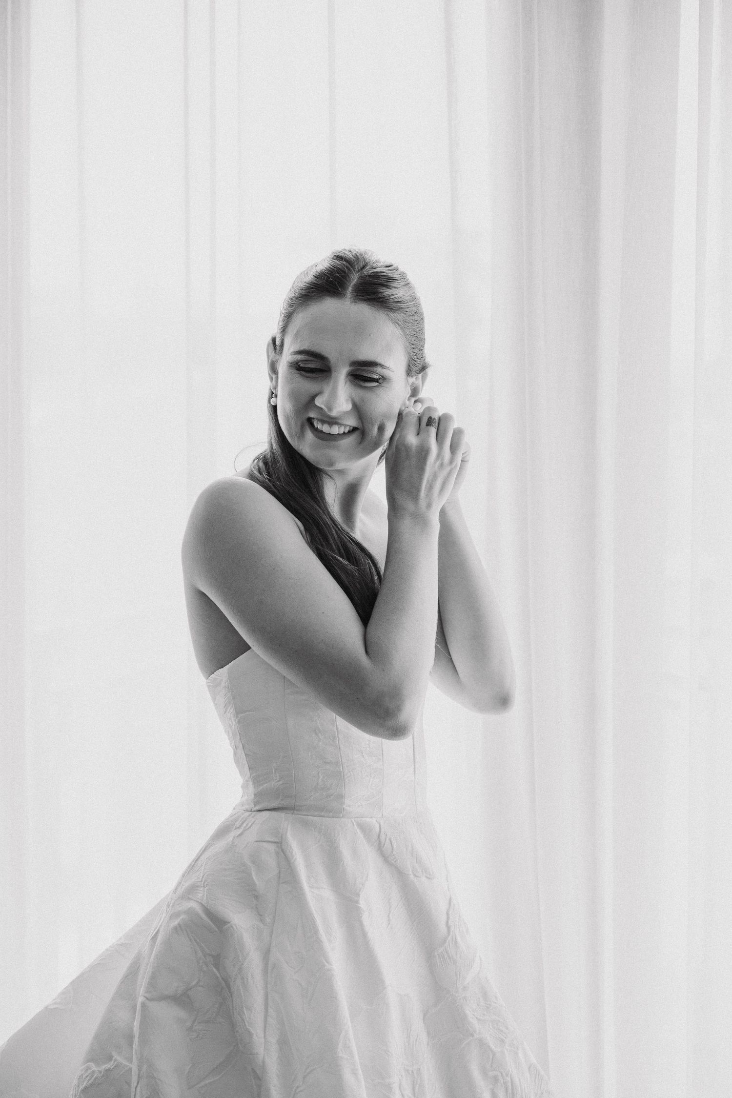 Black and white portrait of someone in a white wedding dress laughing joyfully by a window.