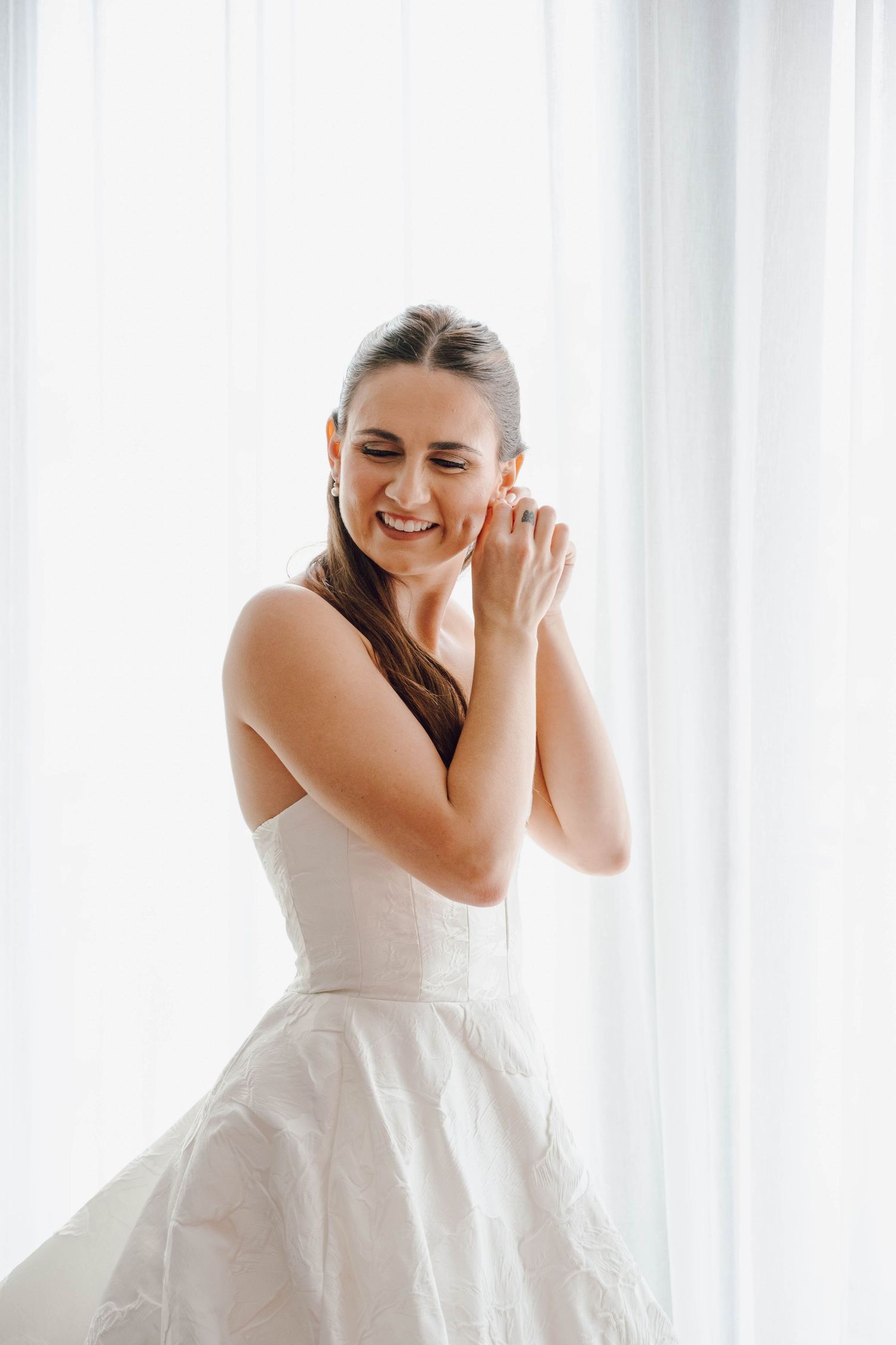 A bride in a white wedding dress laughs joyfully near a bright window in a candid moment.
