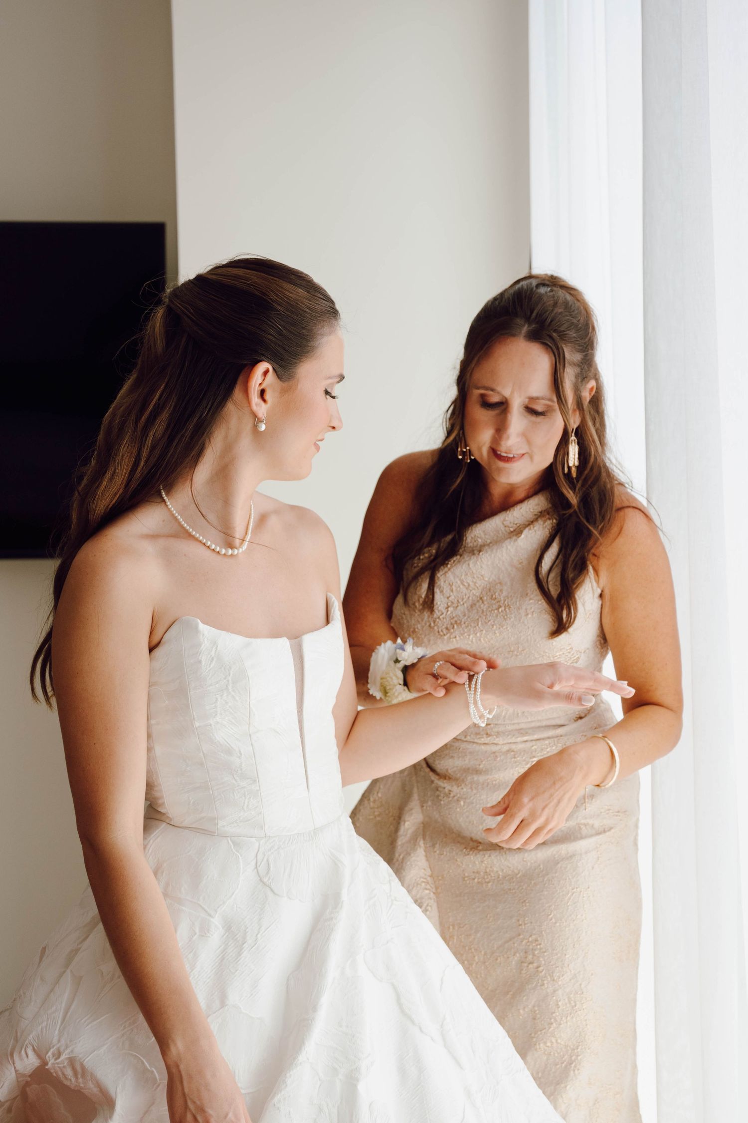 Two people in white dresses share an intimate moment while looking down at something together in a bright room.