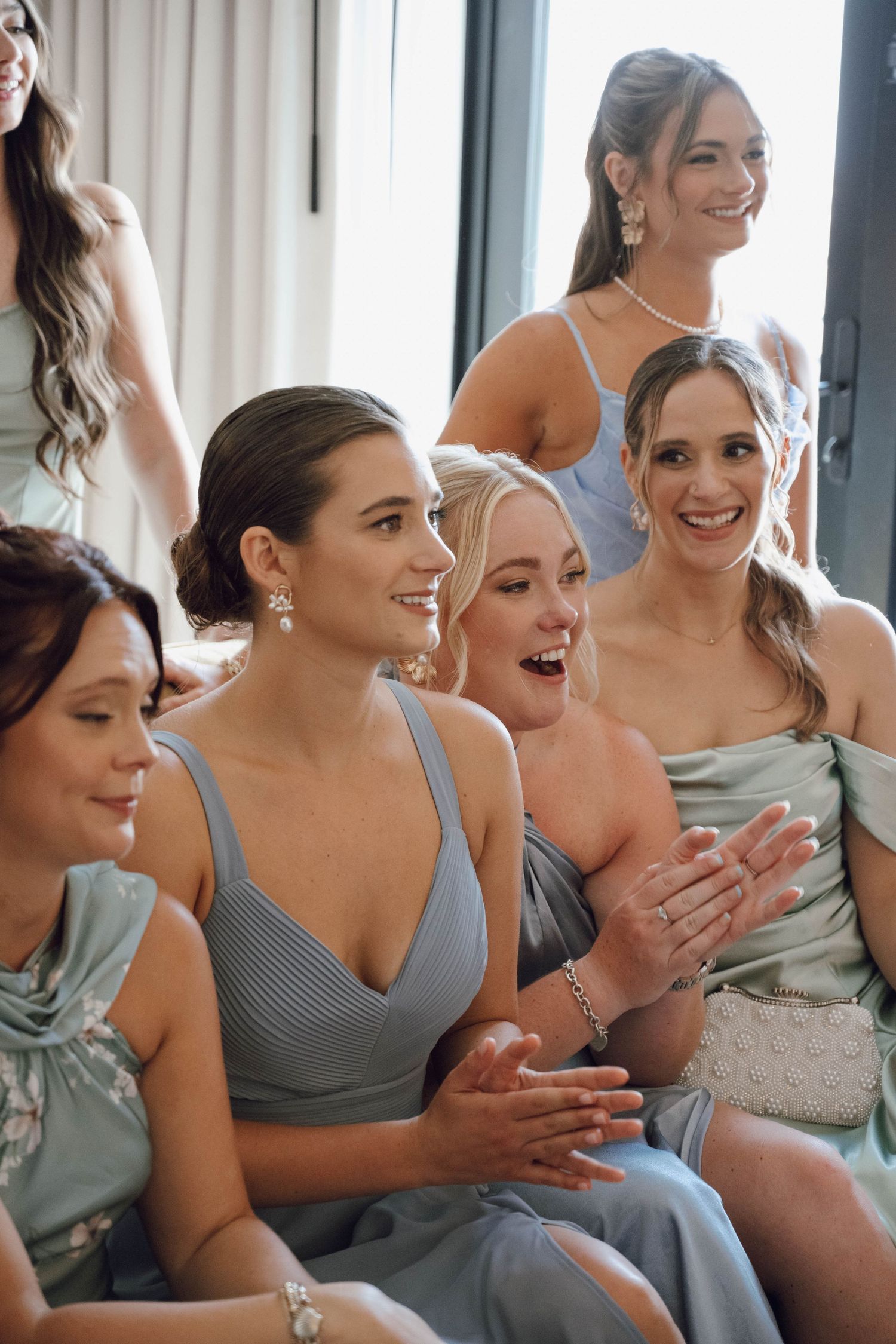 Bridesmaids in sage green dresses smile and laugh together during a wedding celebration.
