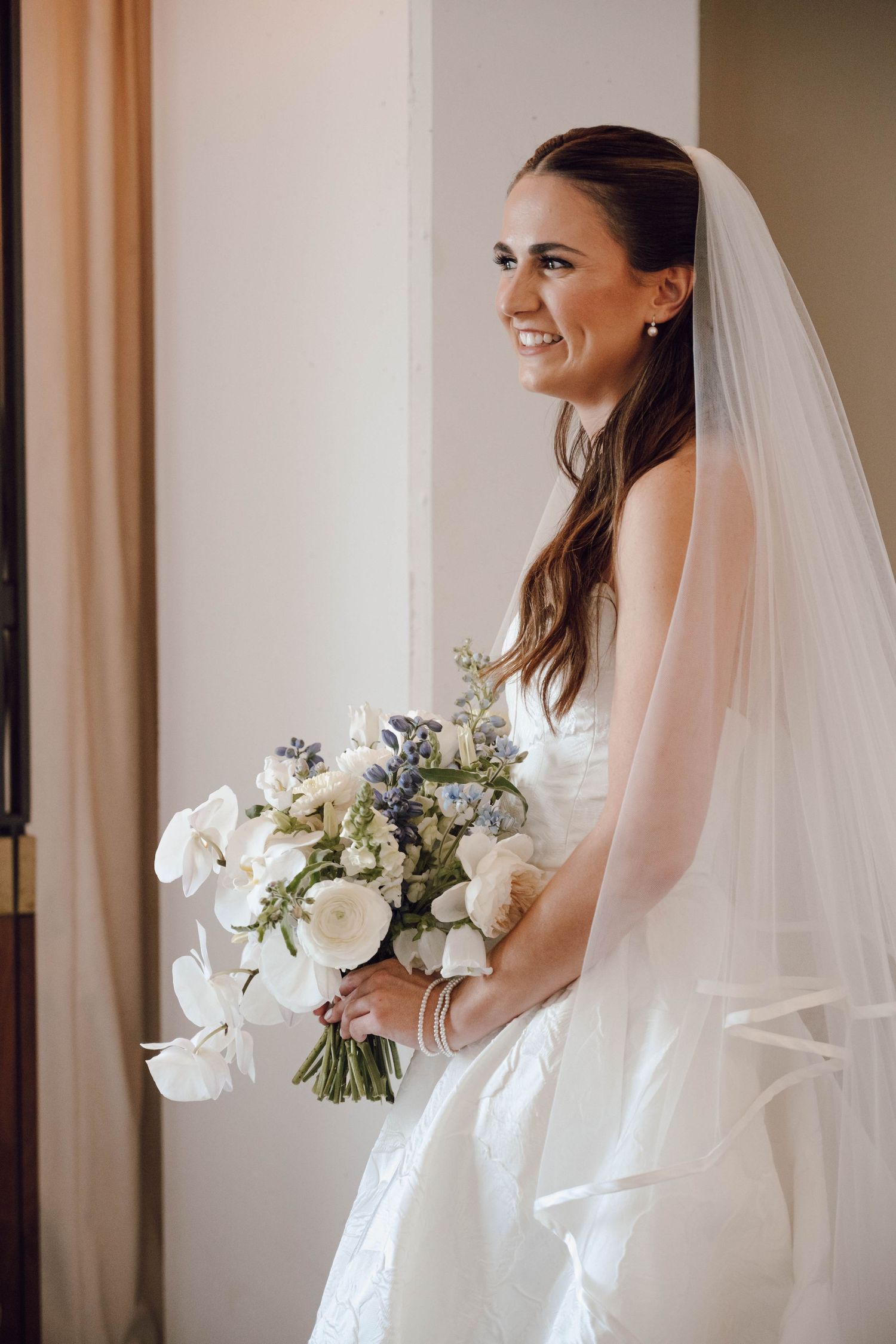 A bride in a white wedding dress and veil holds a bouquet of white flowers against a neutral background.