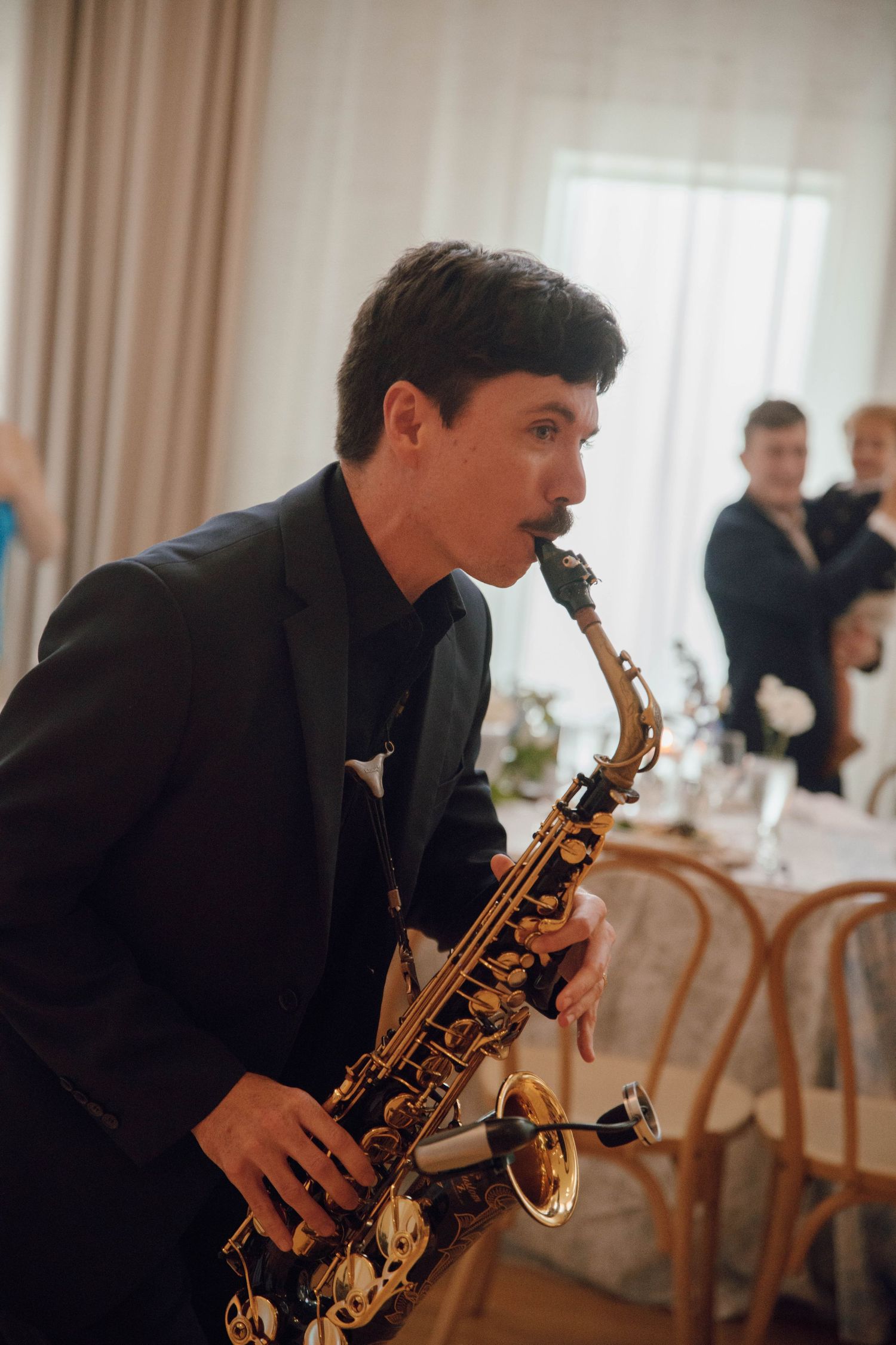 A saxophone player performs at an elegant indoor wedding reception venue with soft lighting and wooden chairs.
